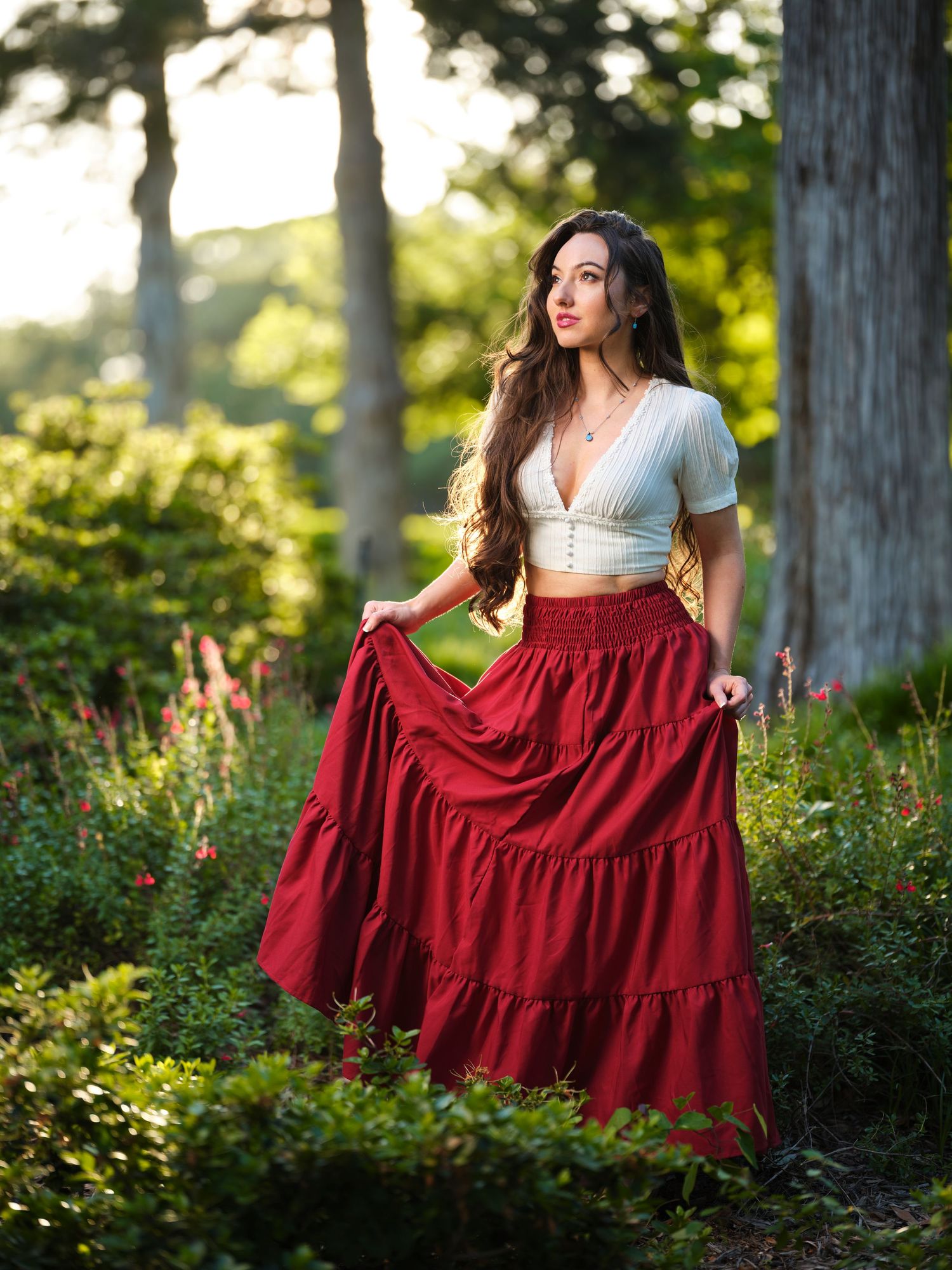 Woman in white crop top and flowing red maxi skirt posing in a sunlit forest.