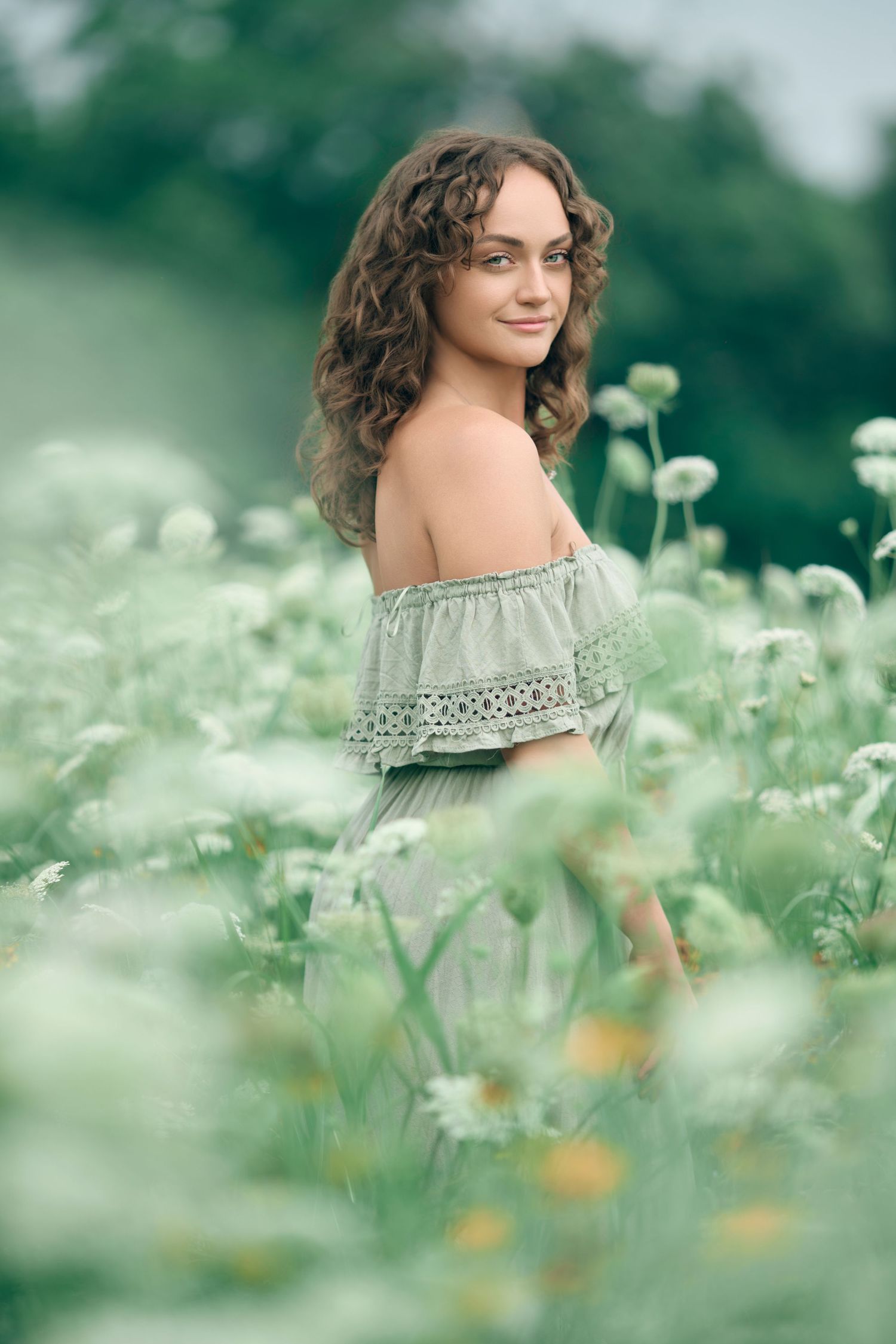 Young woman with curly hair in off-shoulder green dress sitting among white wildflowers in a field.