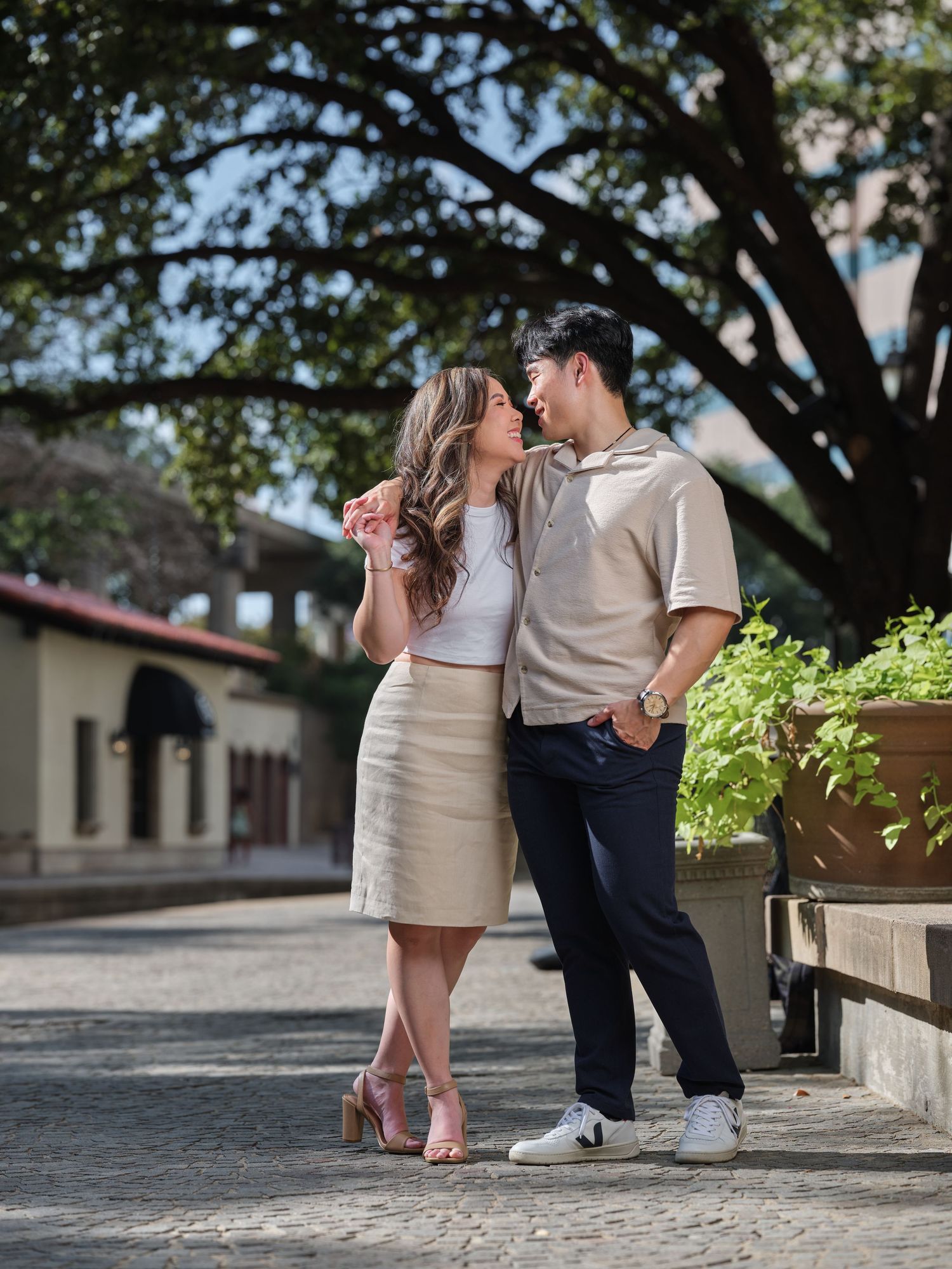 Couple sharing a romantic kiss outdoors on cobblestone path beneath large oak tree in casual attire.