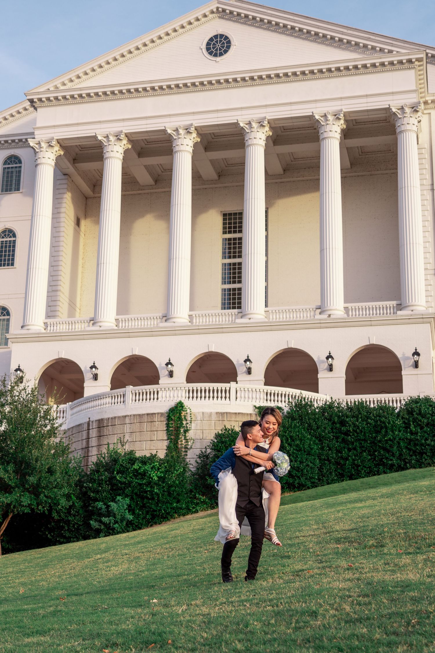 Groom carrying bride in his arms on green lawn in front of grand white columned building.