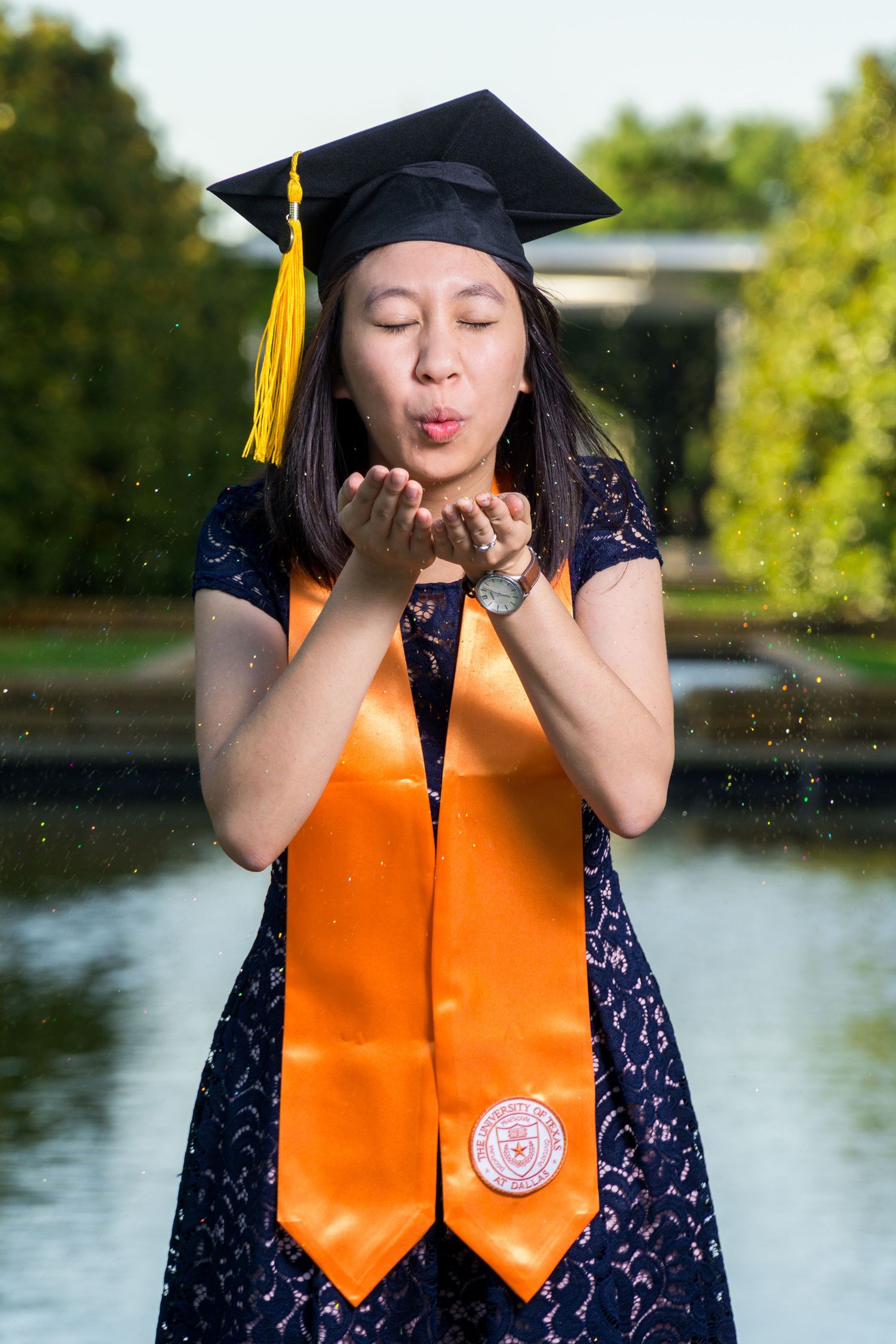 Asian graduate in cap and orange stole blowing glitter from hands near outdoor fountain.