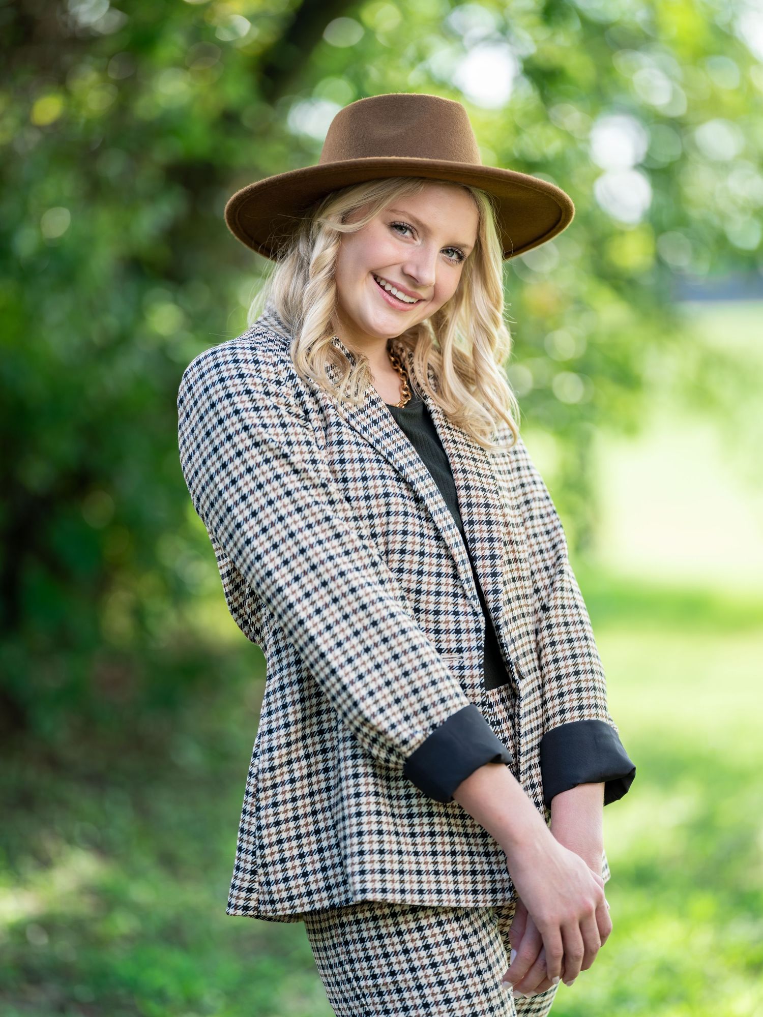 Smiling young blonde woman wearing a brown wide-brim hat and plaid blazer posing outdoors in a garden.