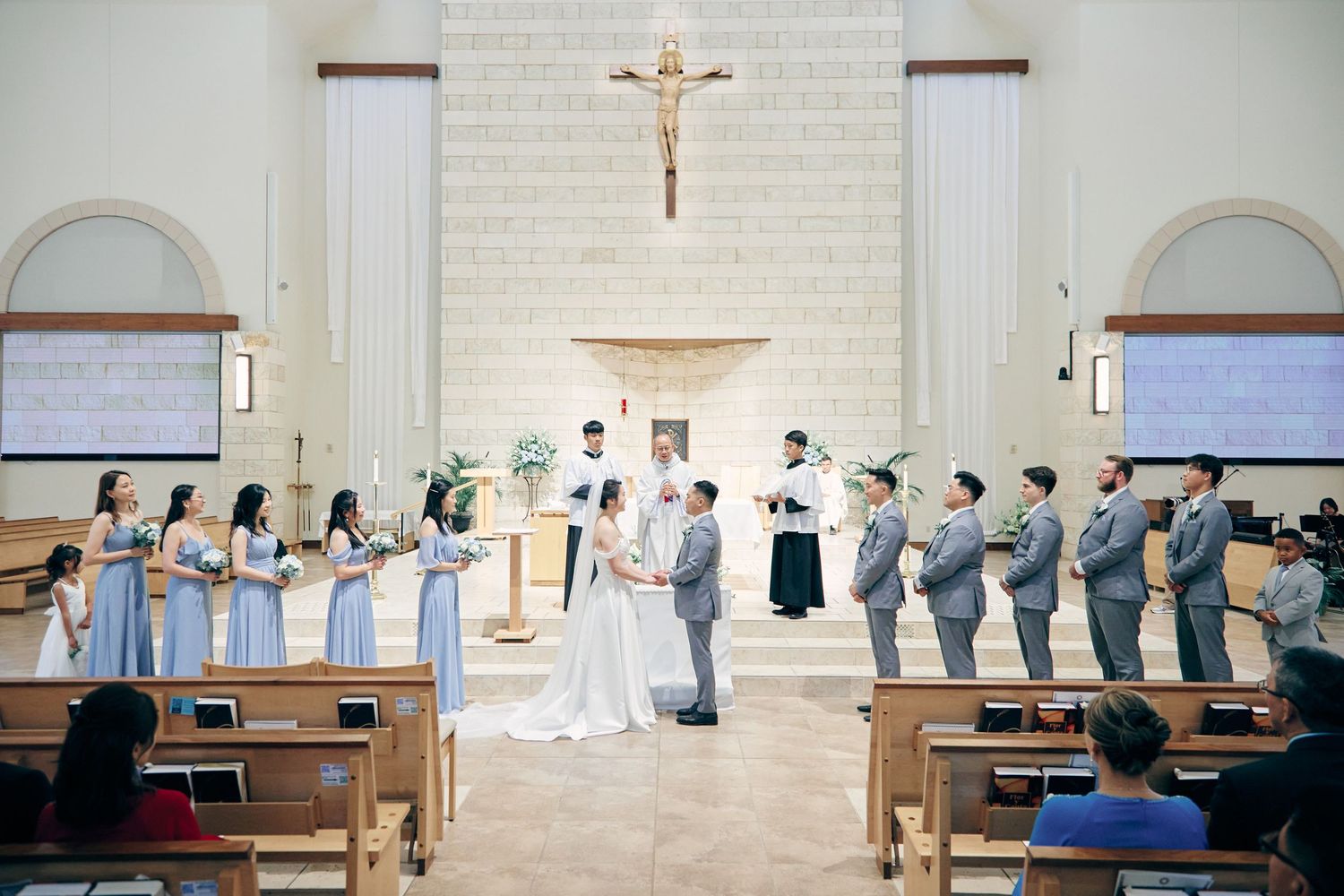 Bride and groom exchange vows at church altar surrounded by bridesmaids in blue and groomsmen in gray.
