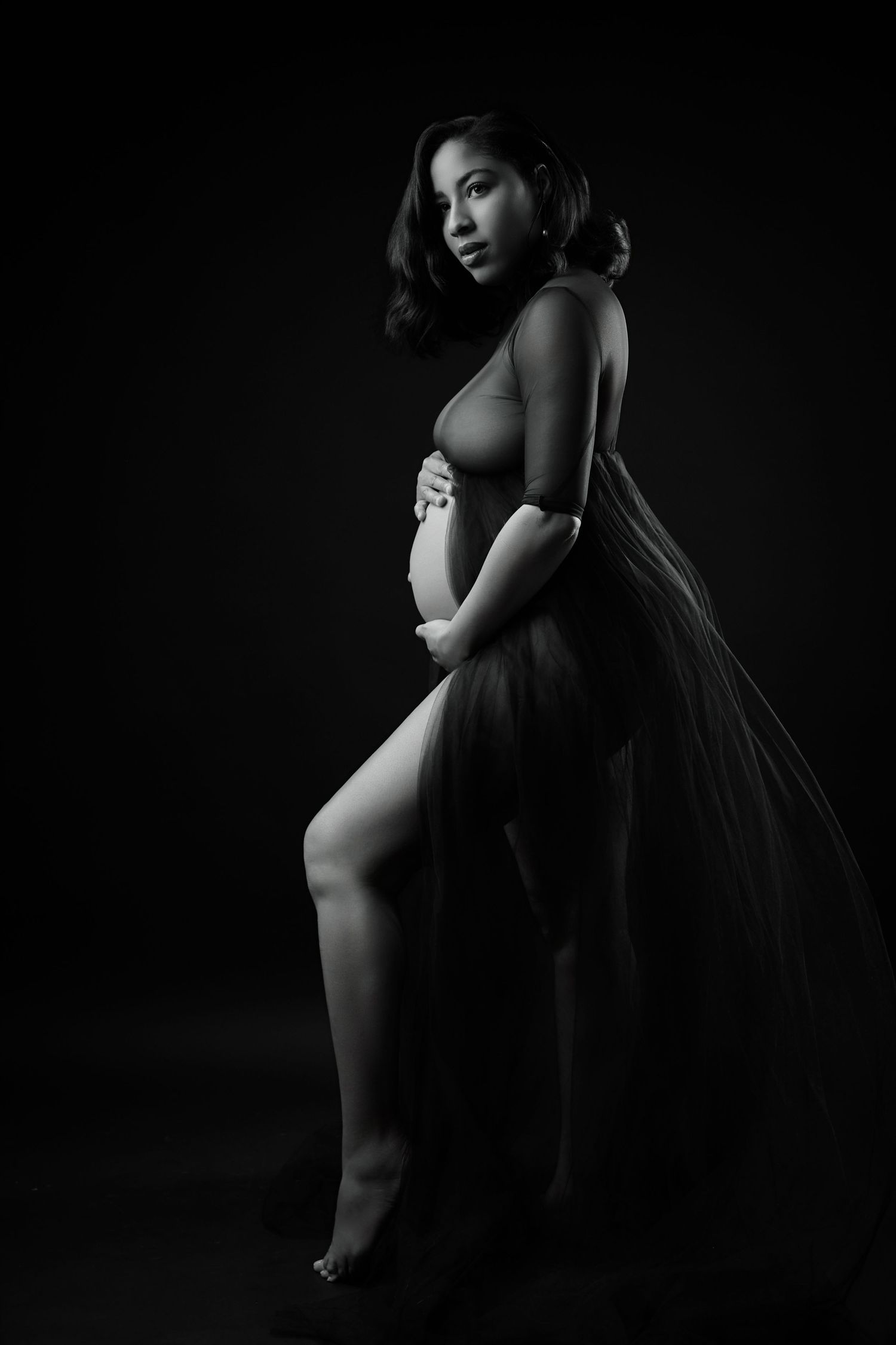 Black and white artistic portrait of pregnant woman in sheer fabric holding her baby bump.