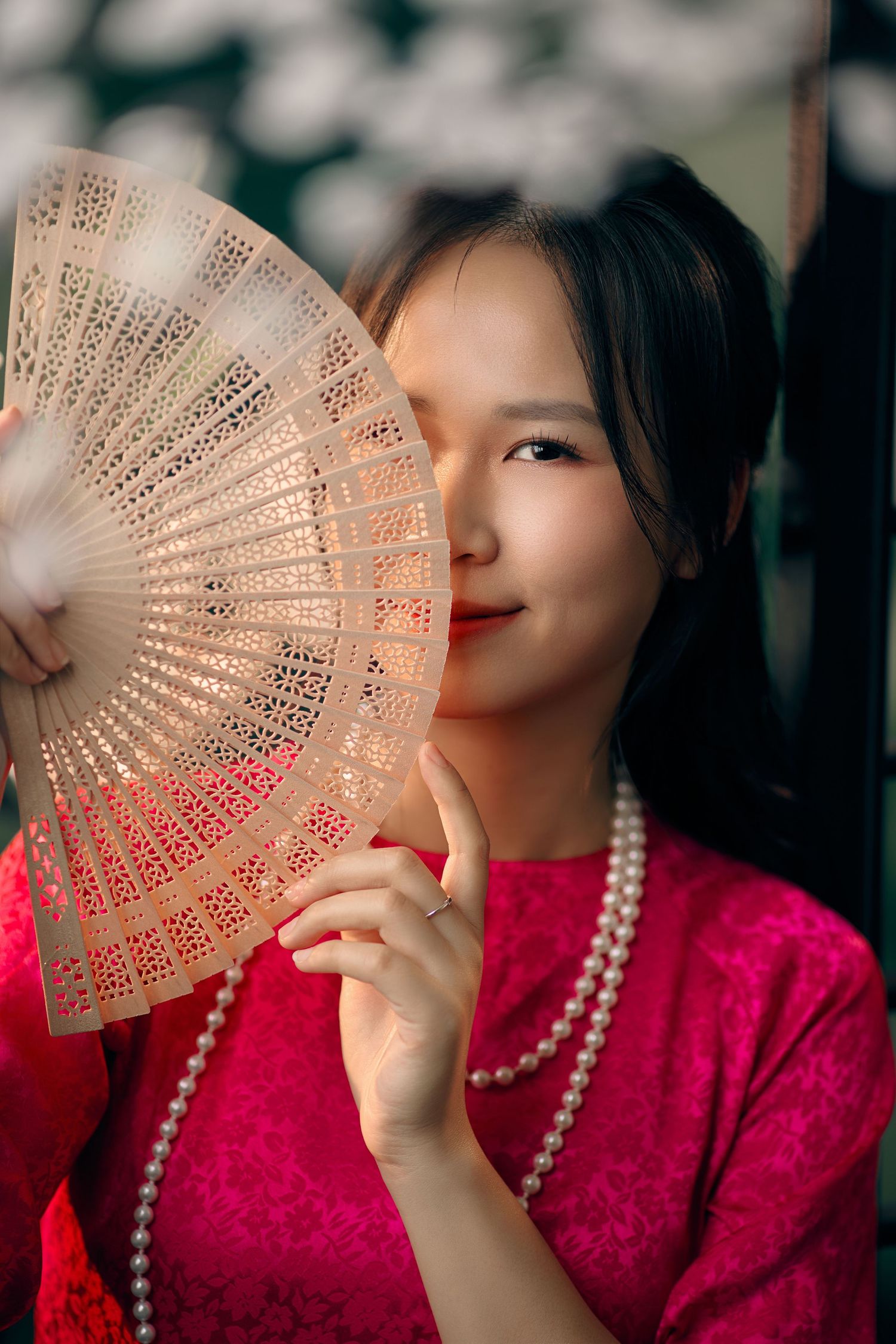 Young woman in pink ao dai holding decorative wooden fan with pearl necklace.