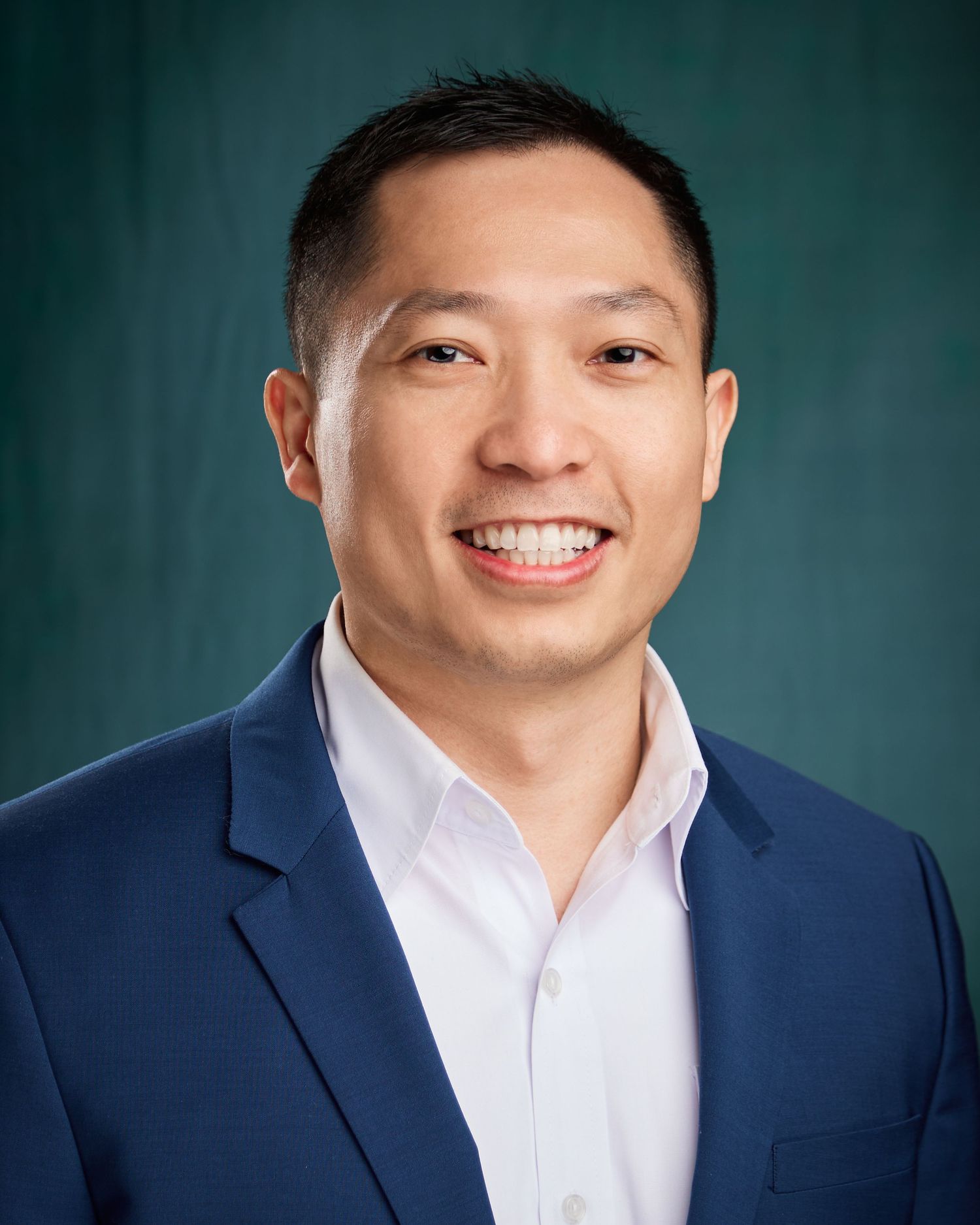 Professional headshot of a smiling Asian man in a blue blazer against a teal background.