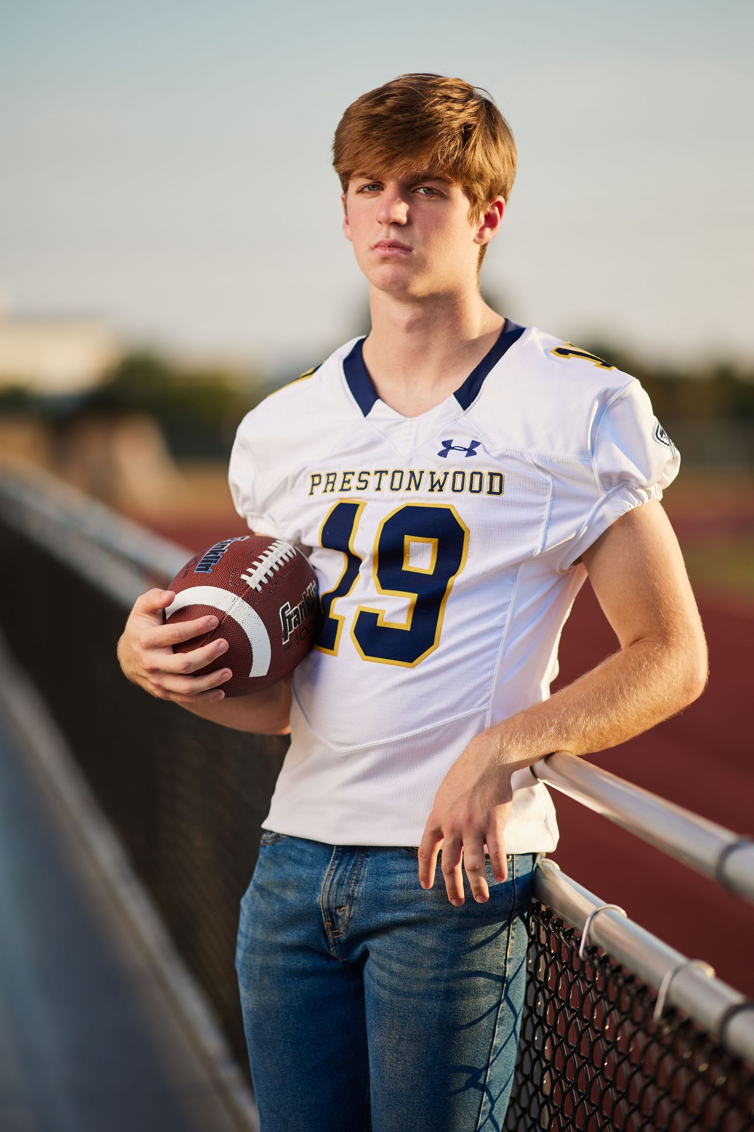 Teen football player in a Prestonwood jersey number 19 holding a football on a high school track.