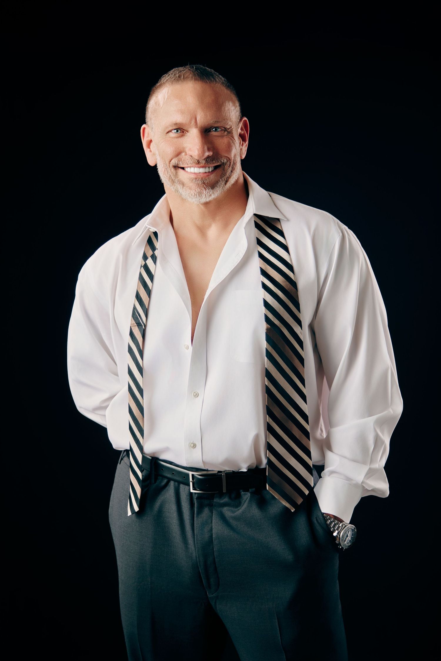 Confident muscular man in white shirt with striped ties smiling against black background.