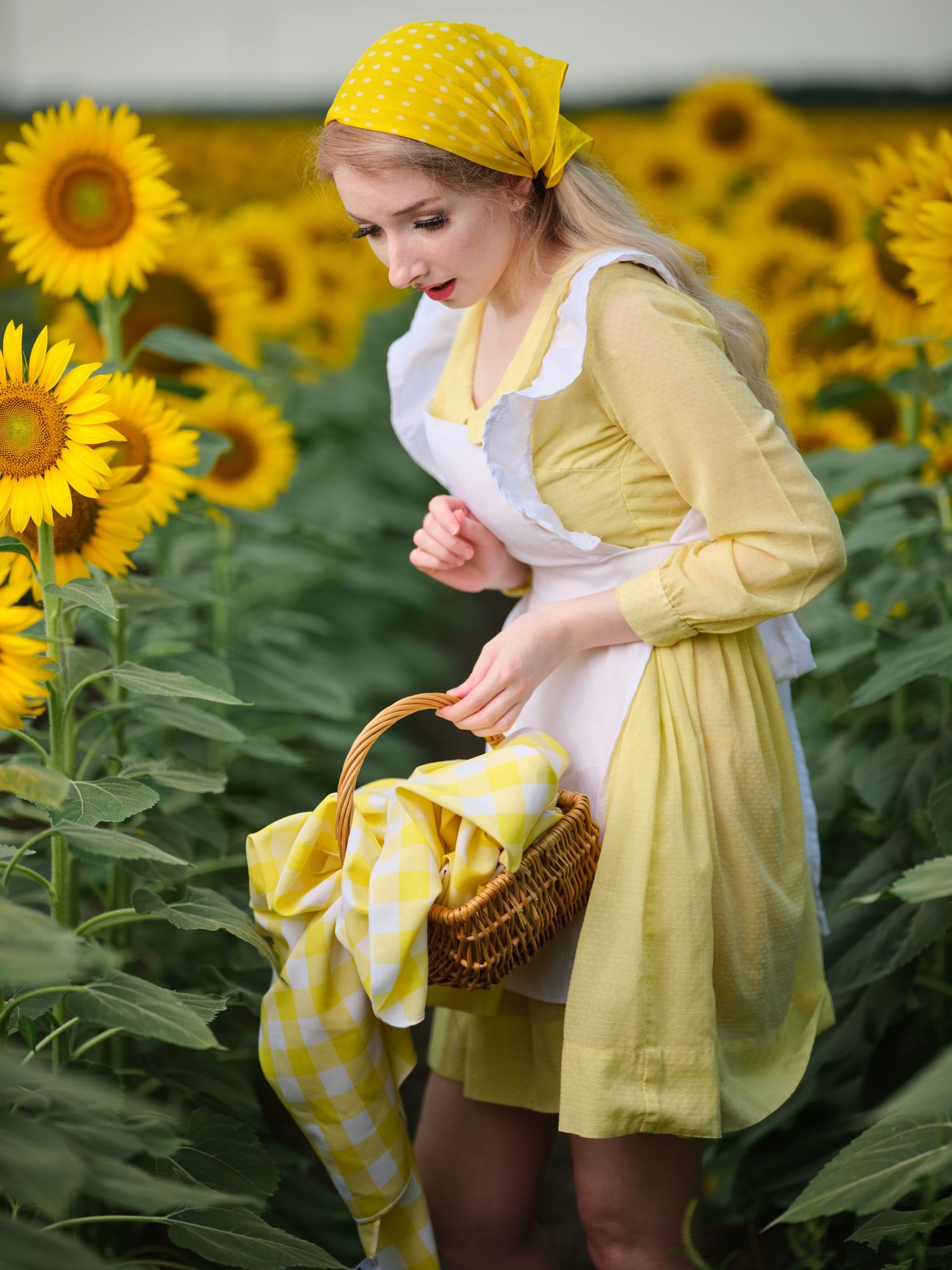 Blonde woman in yellow vintage dress with apron holding basket among sunflowers in a field.
