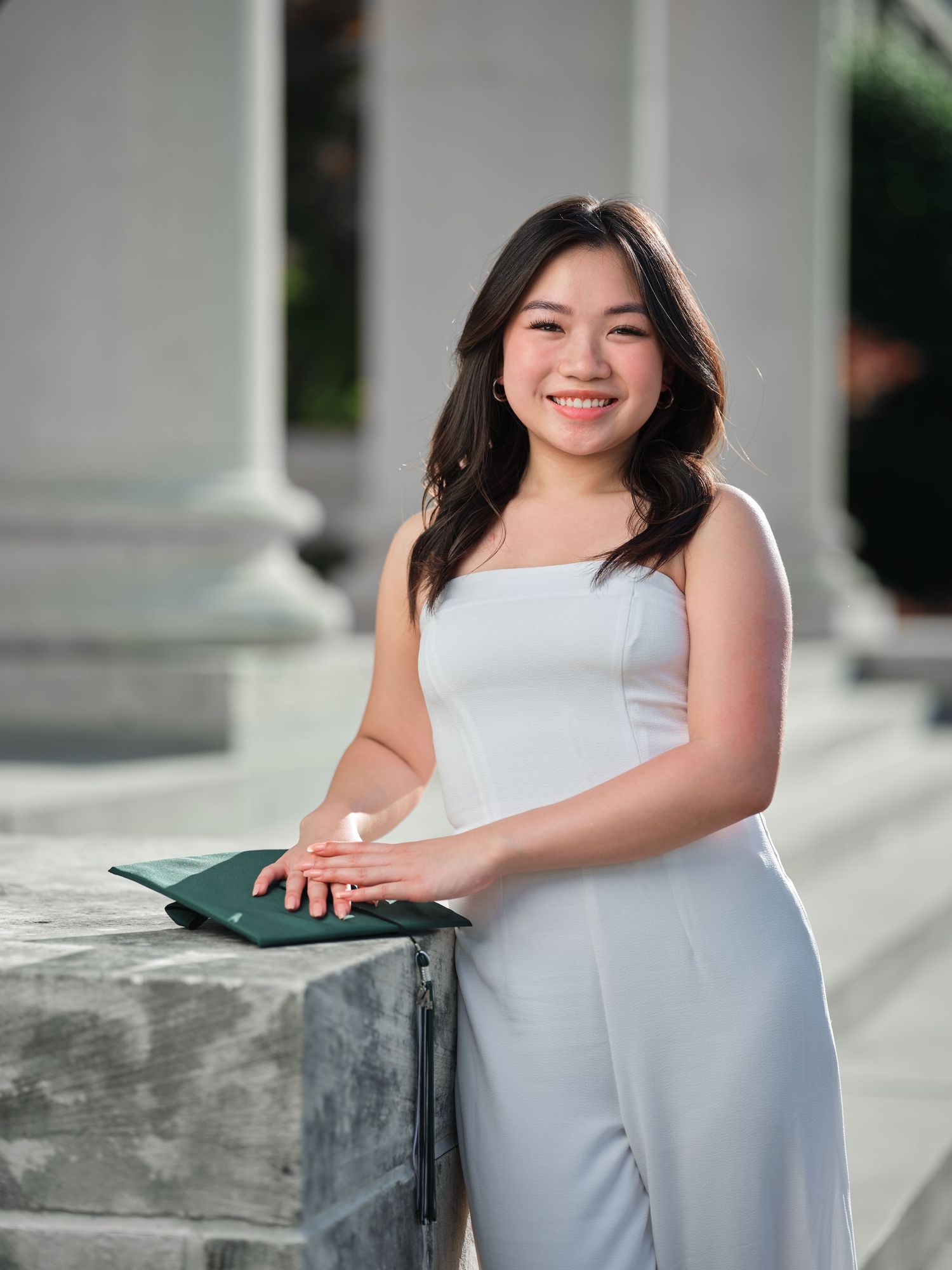 Smiling graduate in white jumpsuit holds green diploma cover while leaning on stone wall.