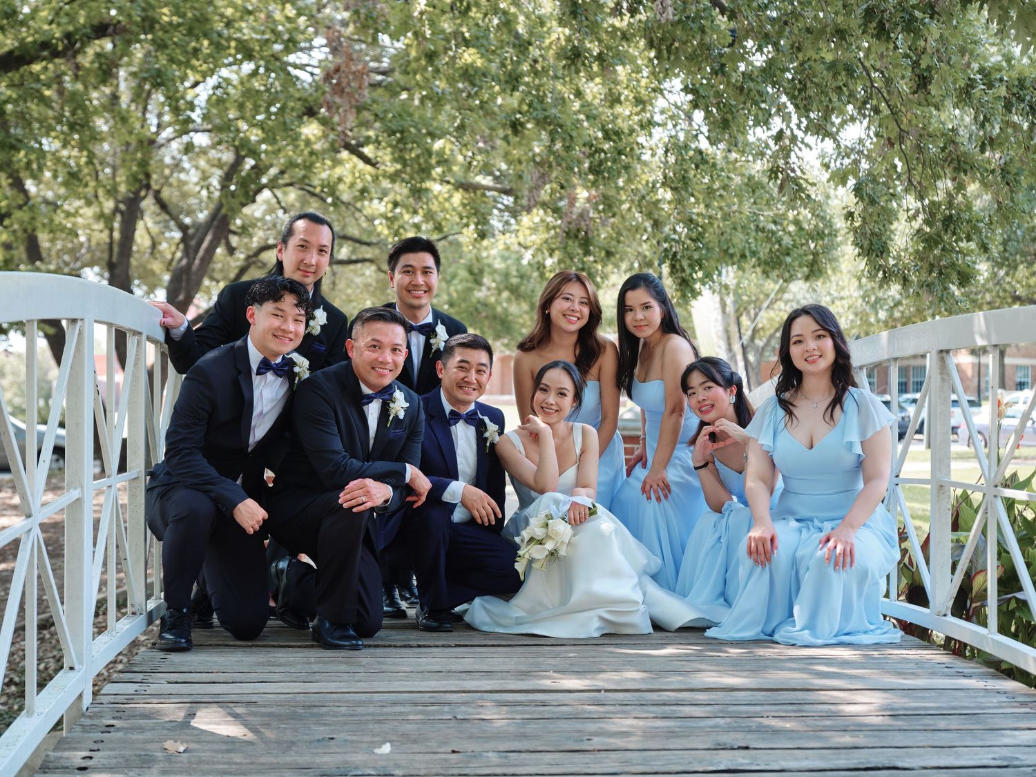 Wedding party group photo on wooden bridge with light blue bridesmaids dresses outdoors.