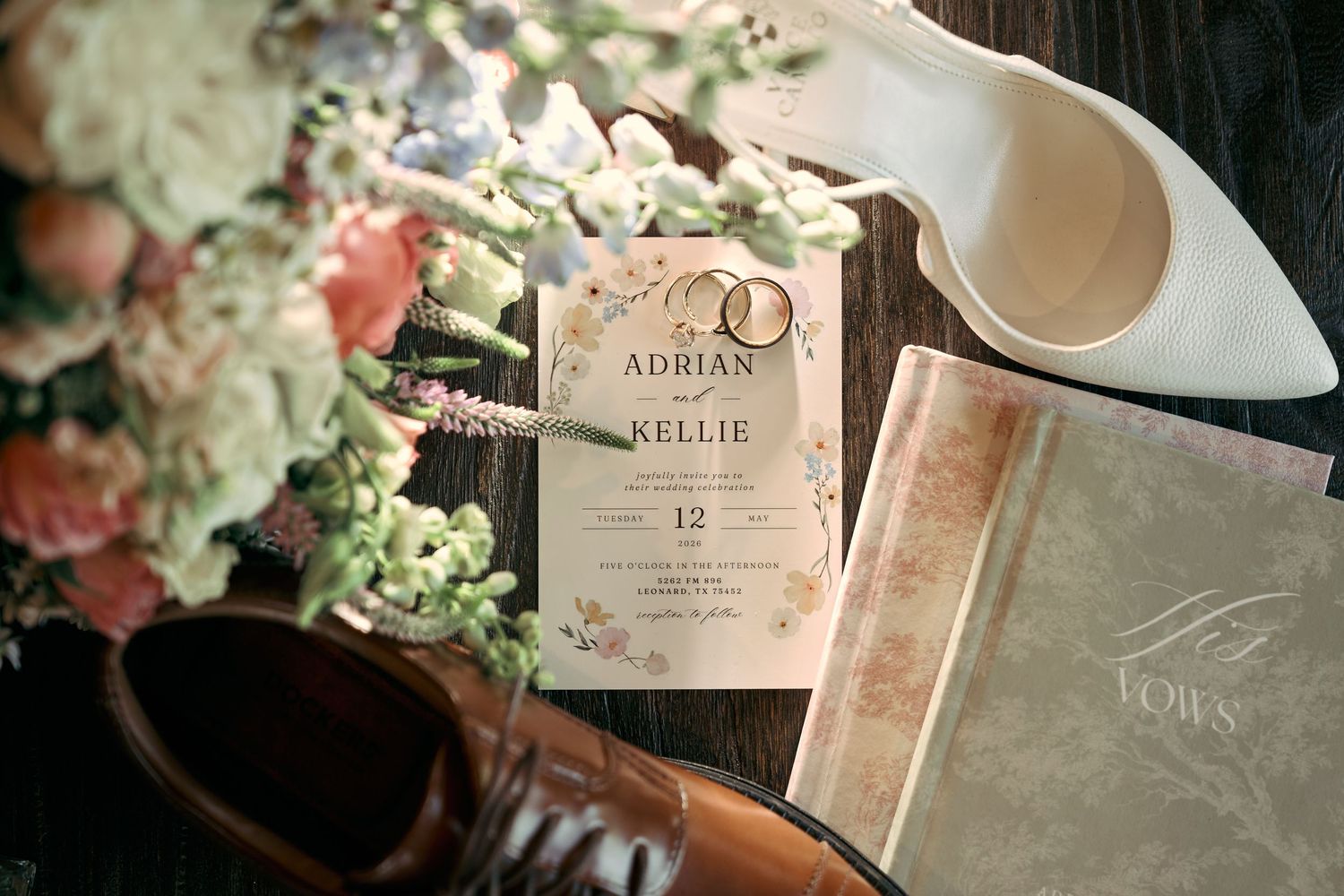 Wedding flat lay featuring Adrian and Kellie invitation, gold rings, bridal heels, bouquet, and brown dress shoes on wooden surface.