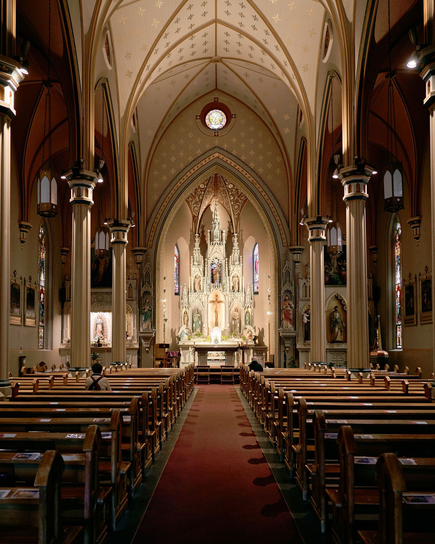 Grand Catholic church interior with ornate Gothic altar, red carpet aisle, wooden pews, and stained glass windows.