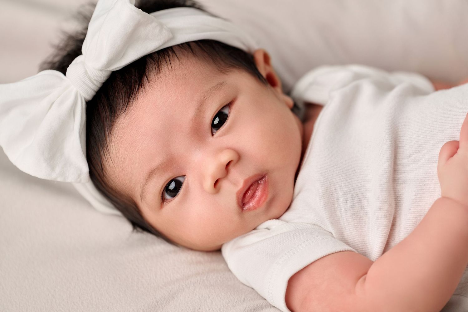 Adorable newborn baby girl wearing a white bow headband and onesie, lying on a soft white blanket.