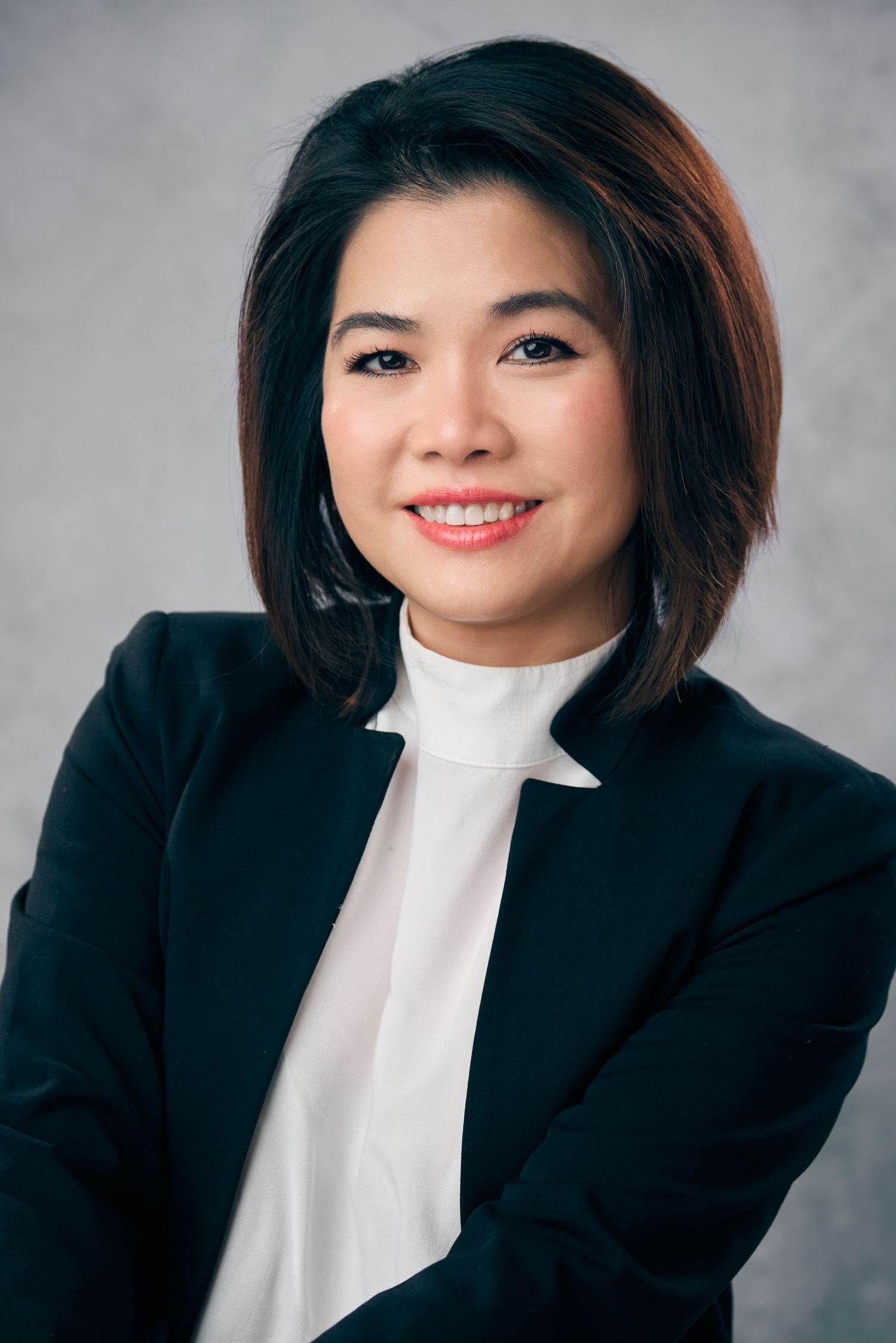 Professional headshot of an Asian woman with bob haircut in a black blazer on gray background.