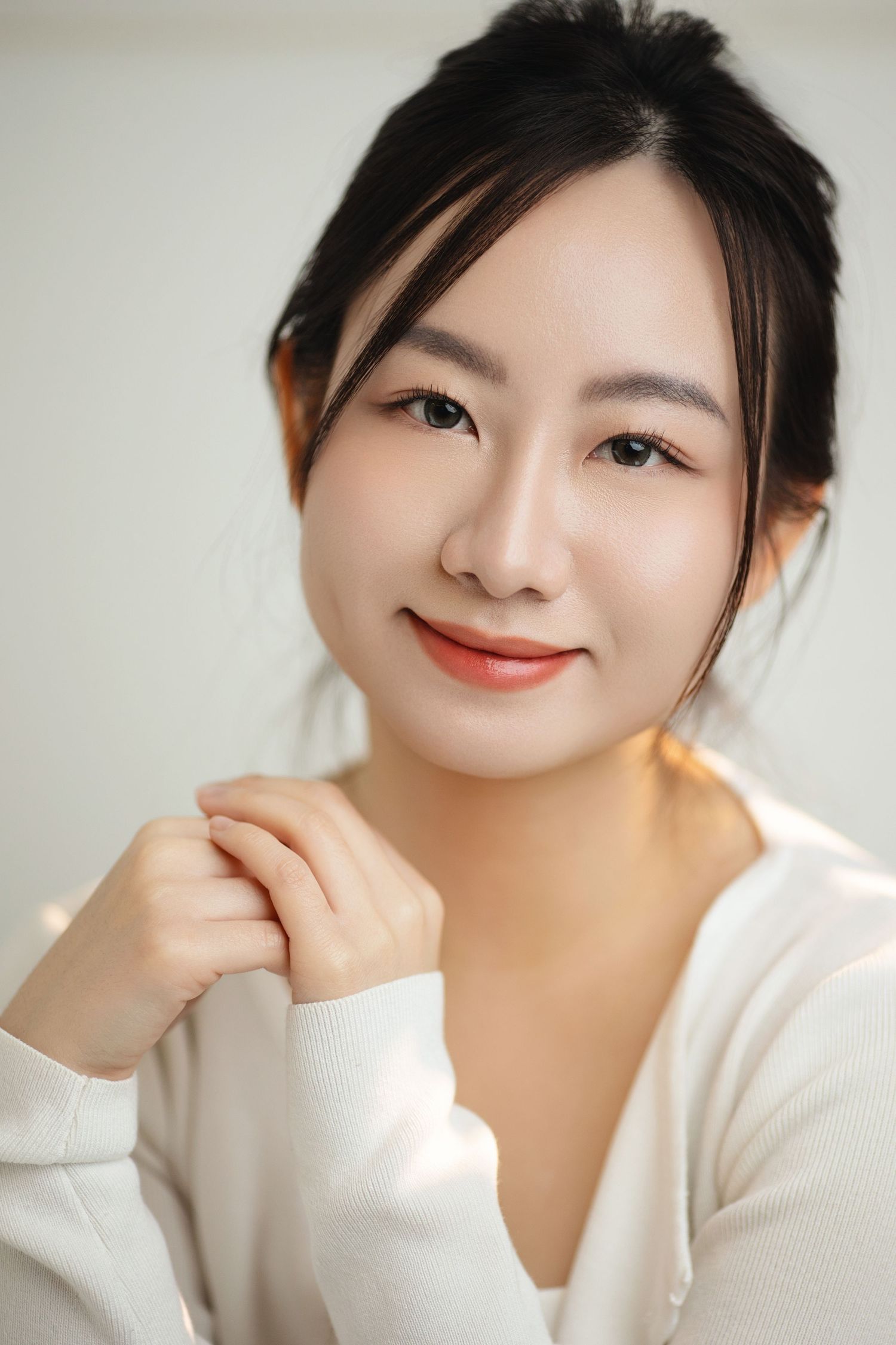 Portrait of a young Asian woman in a white sweater smiling softly against a bright white background.