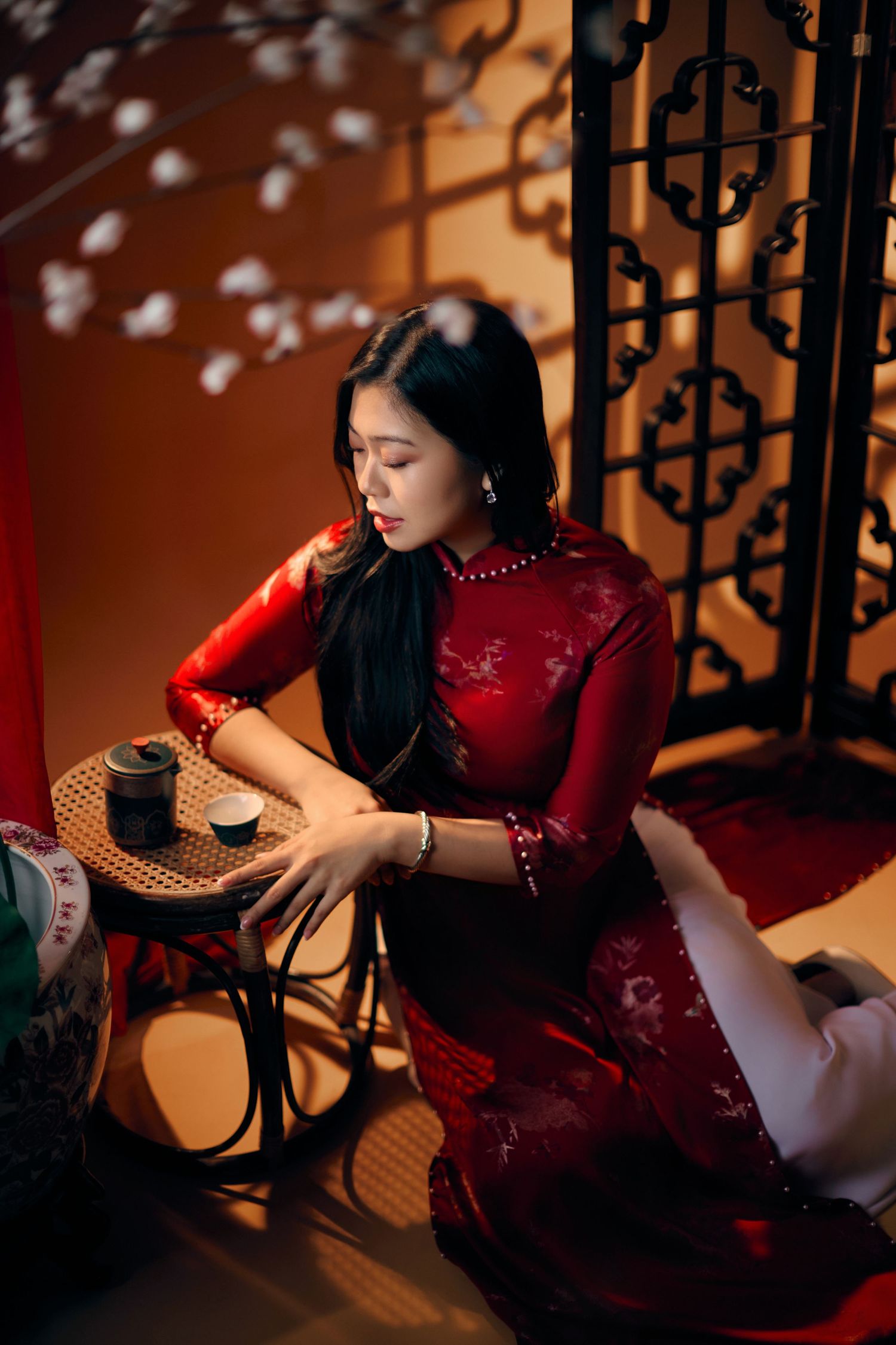 Elegant woman in red floral ao dai sits gracefully beside a small table in warm ambient lighting.