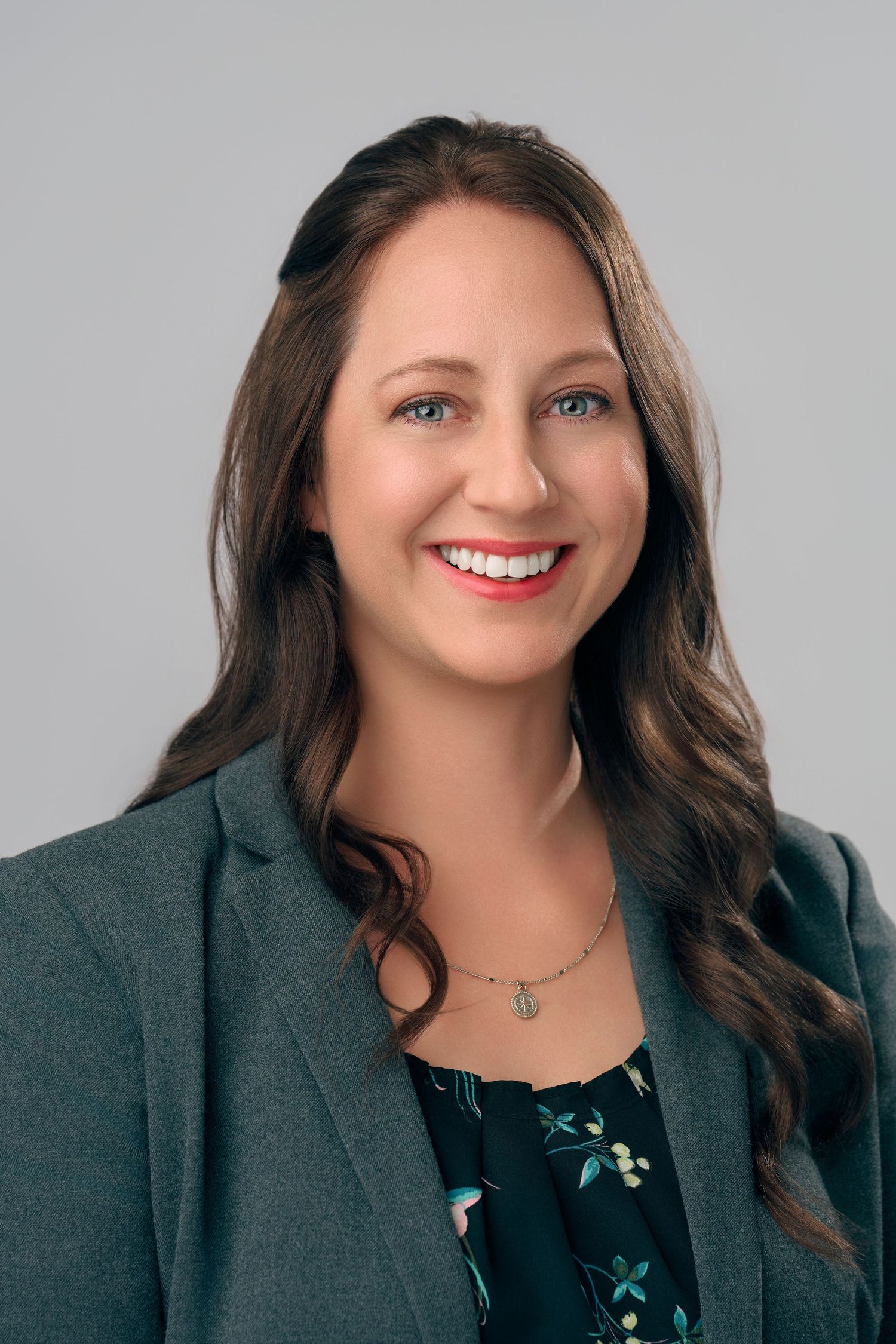 Professional headshot of a smiling woman with brown wavy hair in a gray blazer on gray background.