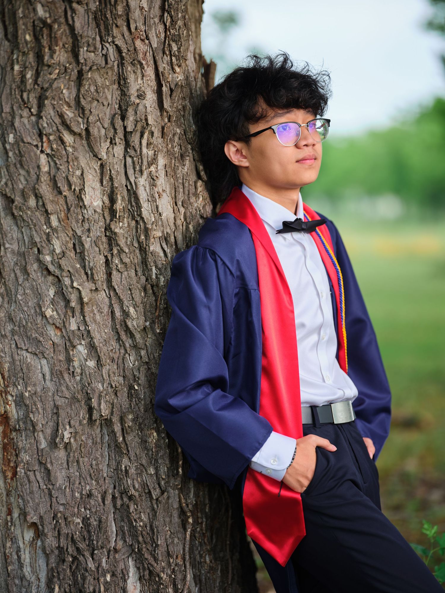 Young man in navy graduation gown and red stole leans against a tree outdoors.