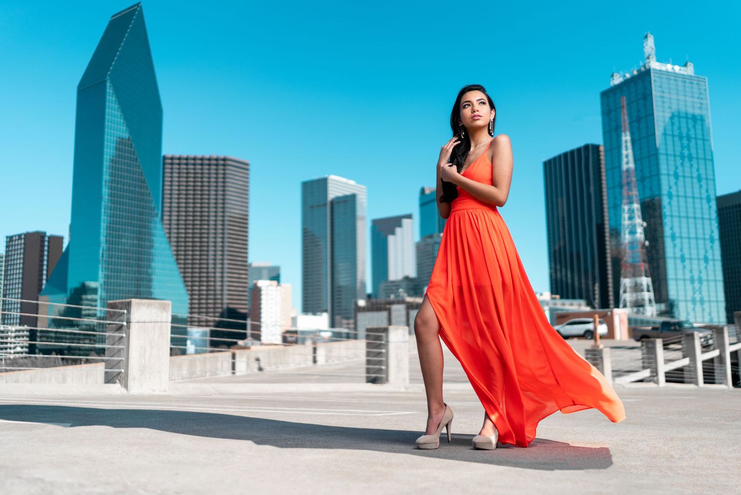 Elegant woman in flowing red dress posing on a rooftop with a dramatic city skyline backdrop.