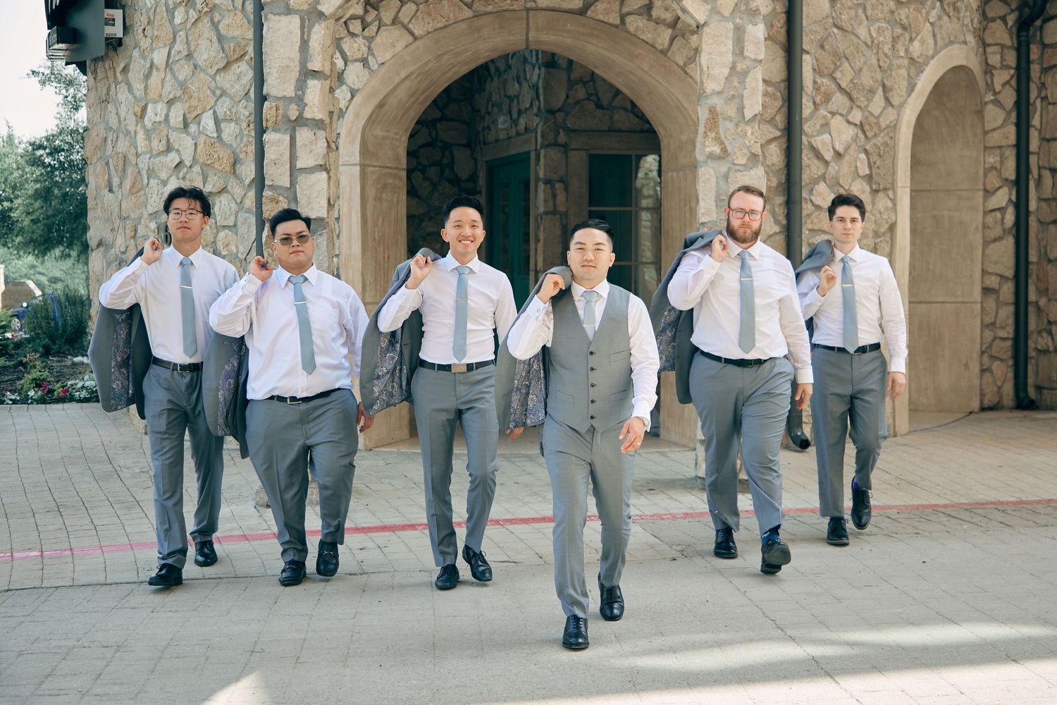 Seven groomsmen in gray suits and ties walking confidently outside a stone church venue.