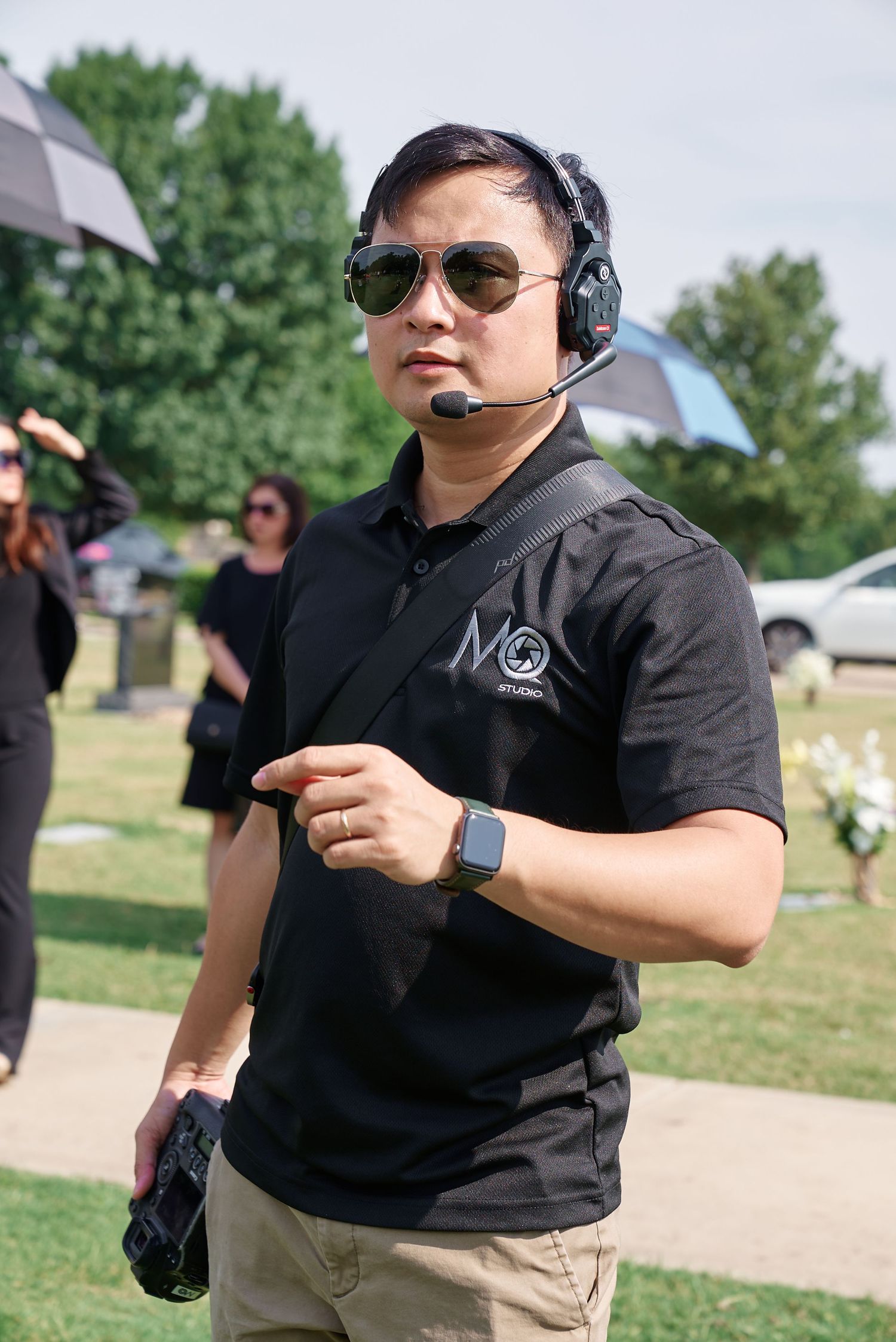 Man in black MQ Studios polo, sunglasses, and headset working outdoors at a cemetery event.