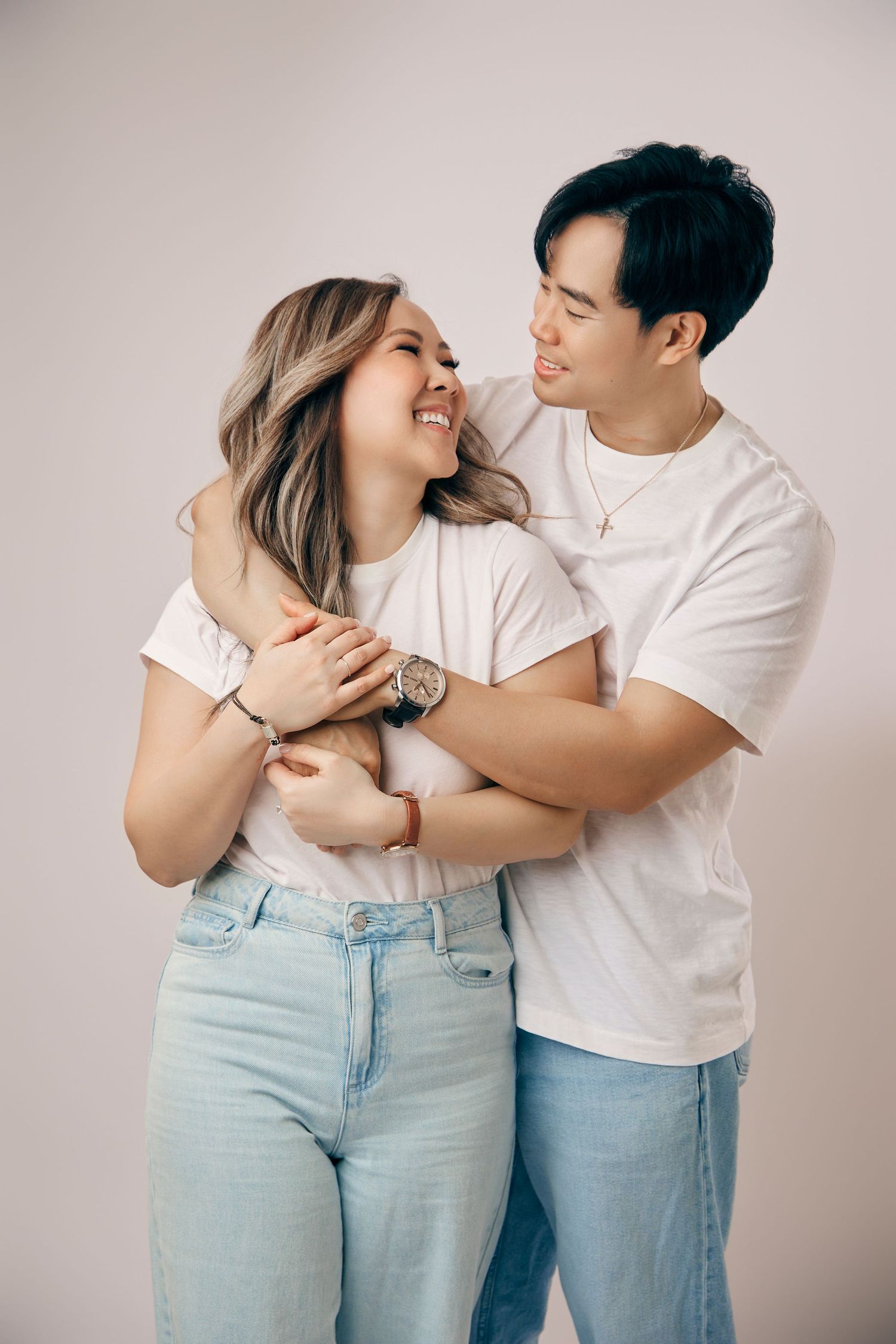 Joyful Asian couple in matching white tees and jeans hugging against a neutral background.