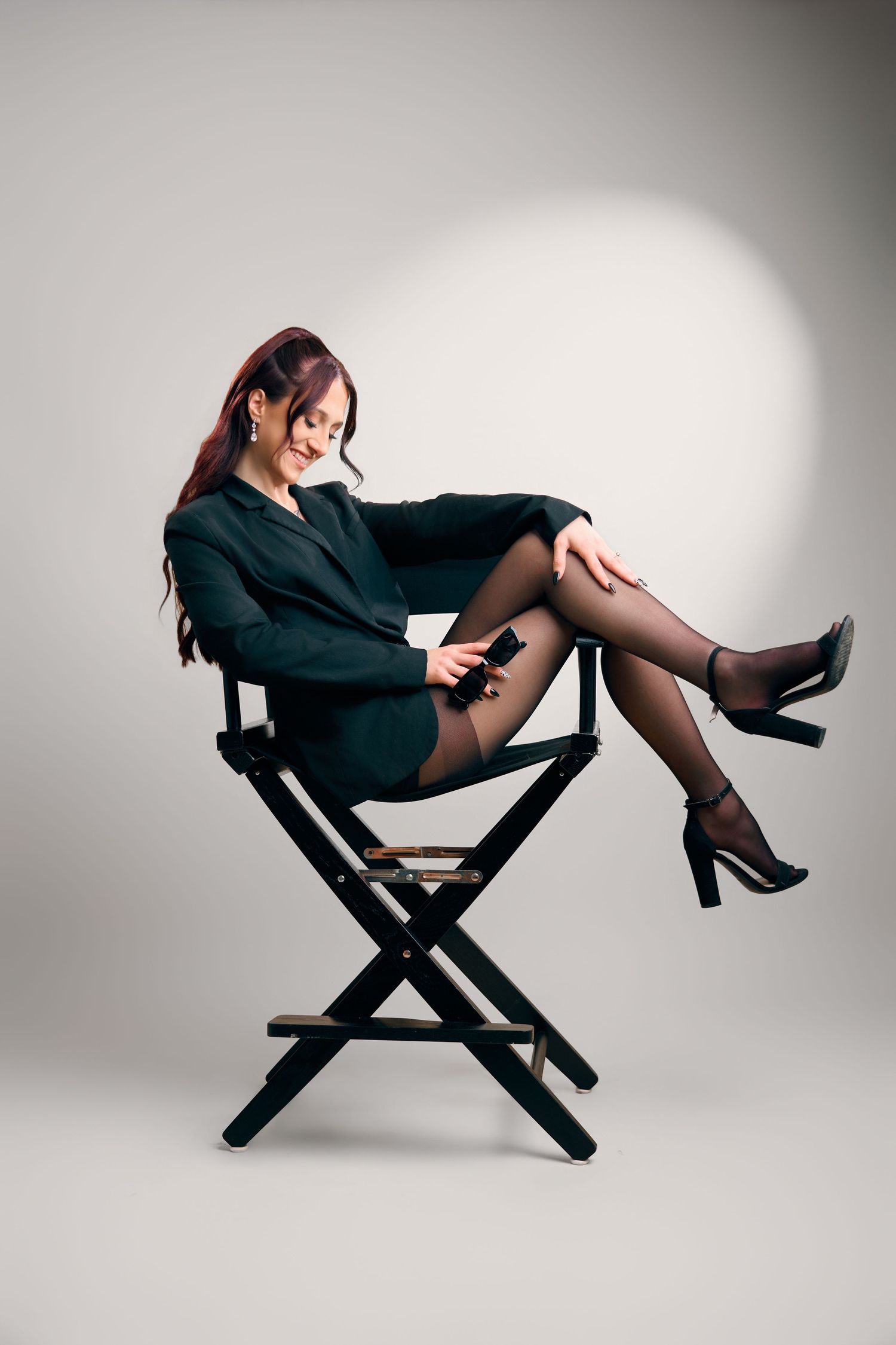 A stylish woman in a black blazer and skirt sits elegantly on a director's chair wearing black stockings and heels.