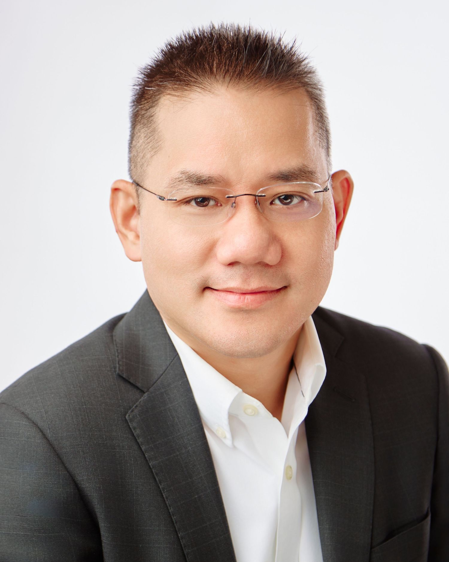 Professional headshot of a smiling Asian man wearing glasses and a gray blazer against a white background.