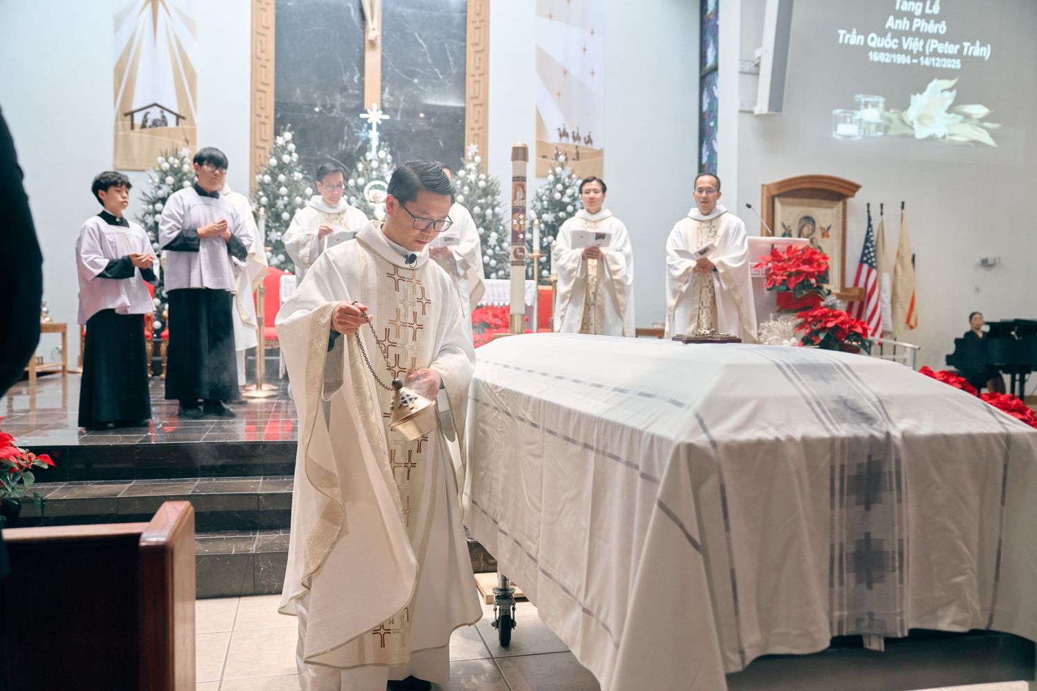 Catholic priest conducting funeral Mass with incense over a white-draped casket inside a decorated church.