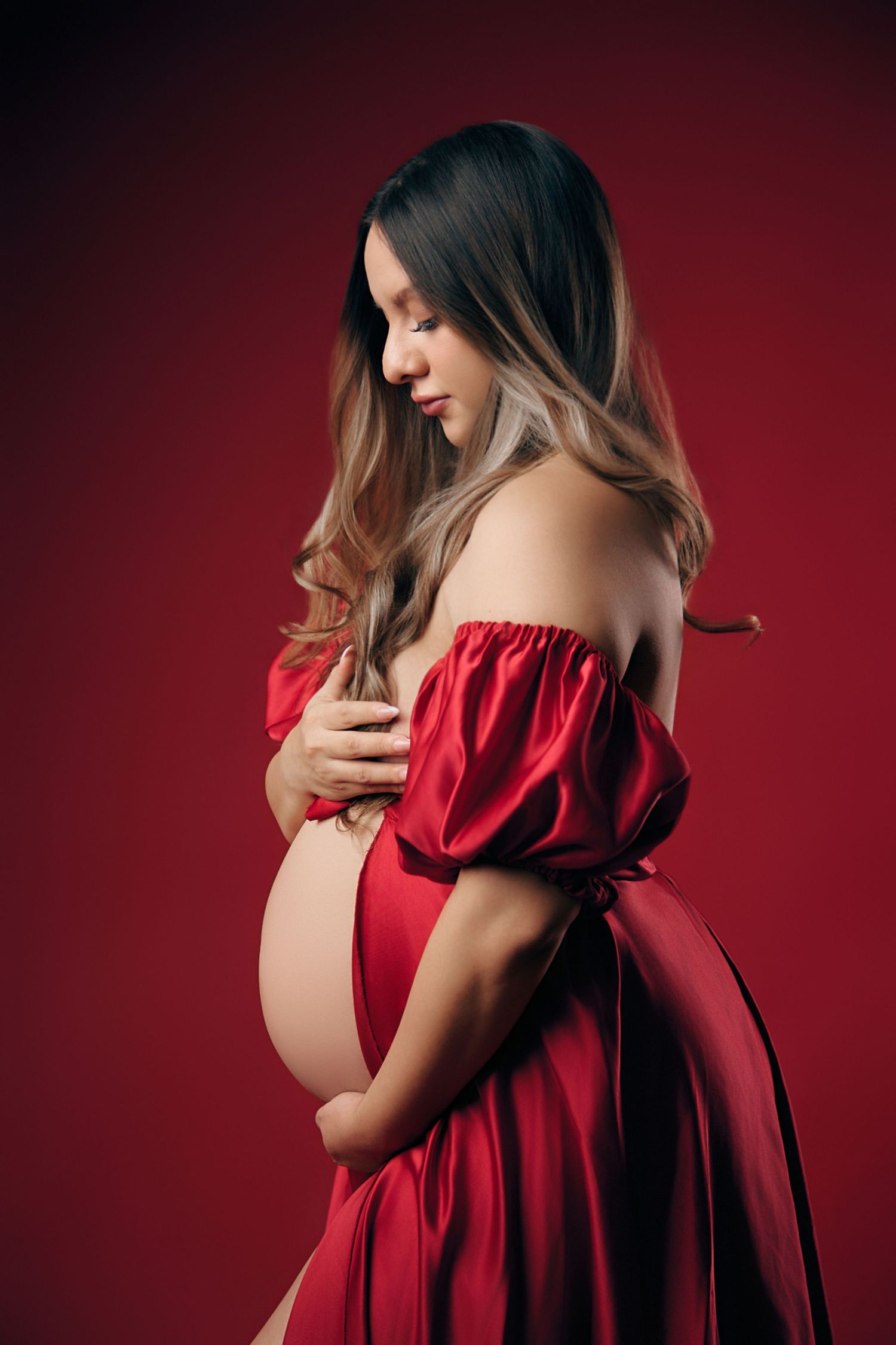 Pregnant woman in elegant red satin off-shoulder gown cradling baby bump on red background.