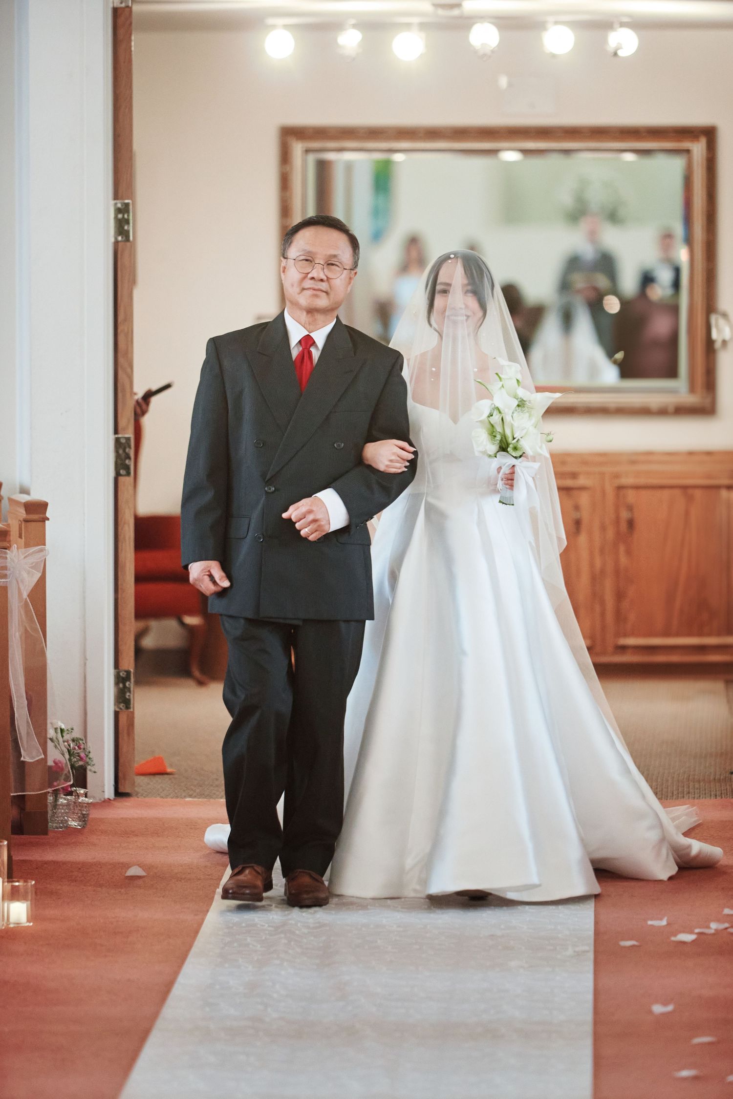 Father walking bride down church aisle in elegant white wedding gown with veil and bouquet.