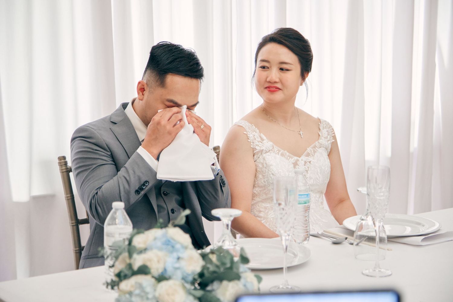 Emotional groom wipes tears while bride smiles warmly during heartfelt wedding reception toasts.