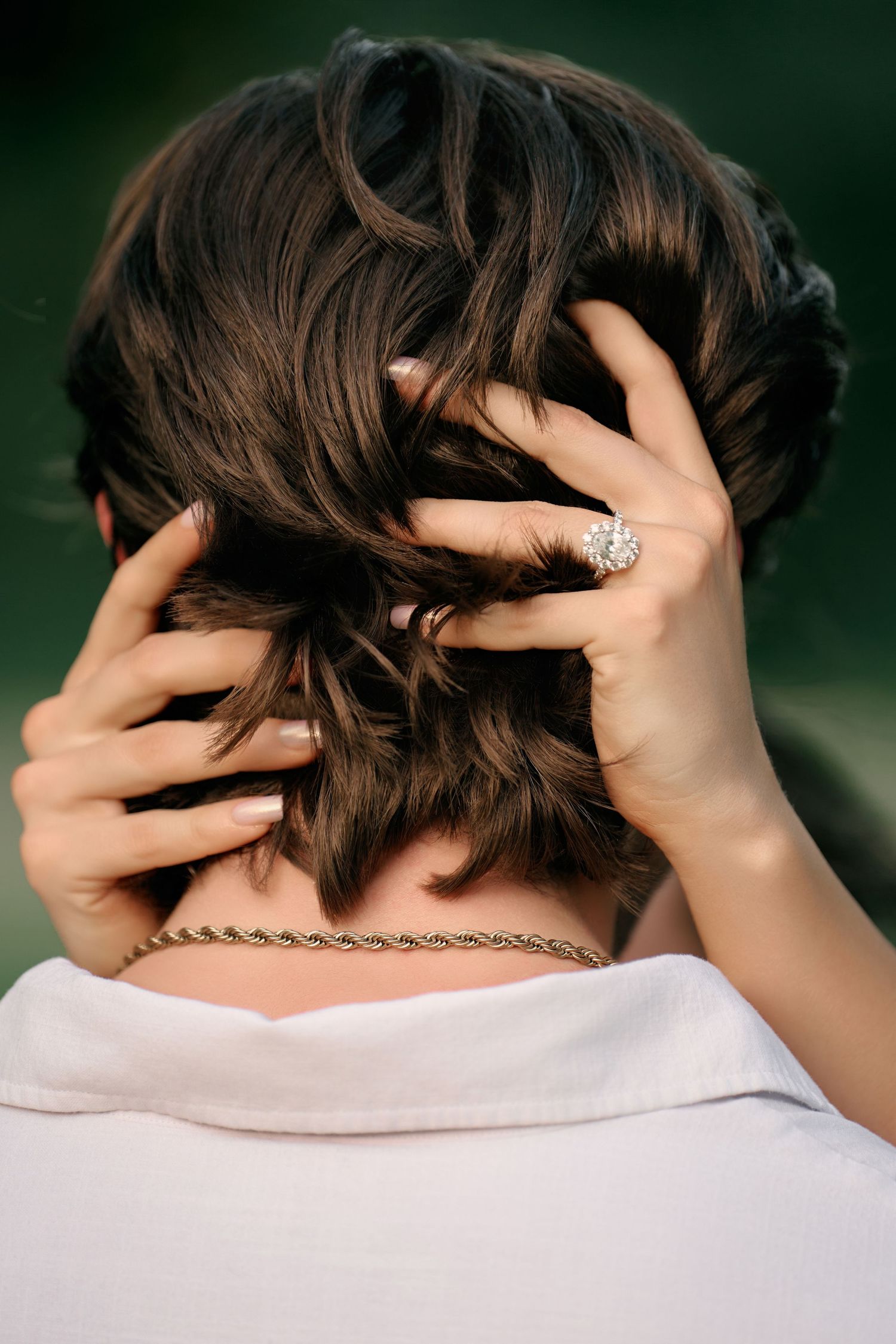 Back view of a person with short dark hair wearing a diamond ring and gold chain necklace.