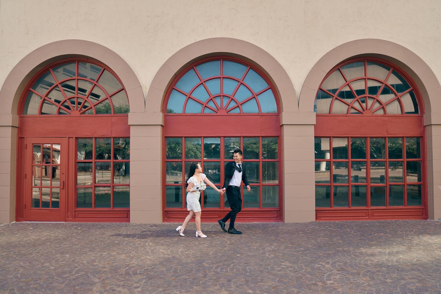 Couple holding hands dancing in front of red arched windows and doors on a beige building exterior.