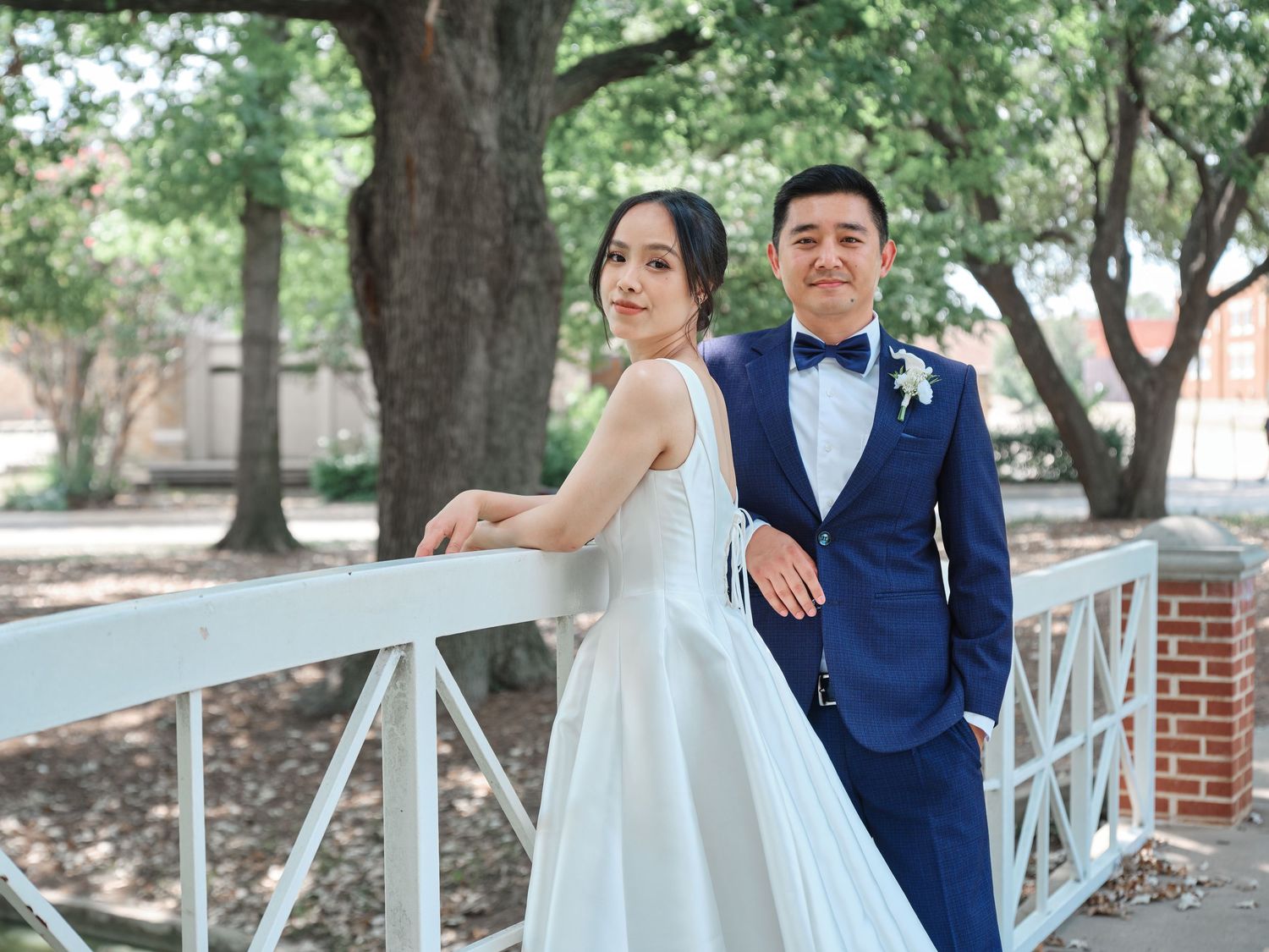 Bride and groom pose together on a white bridge outdoors surrounded by trees.