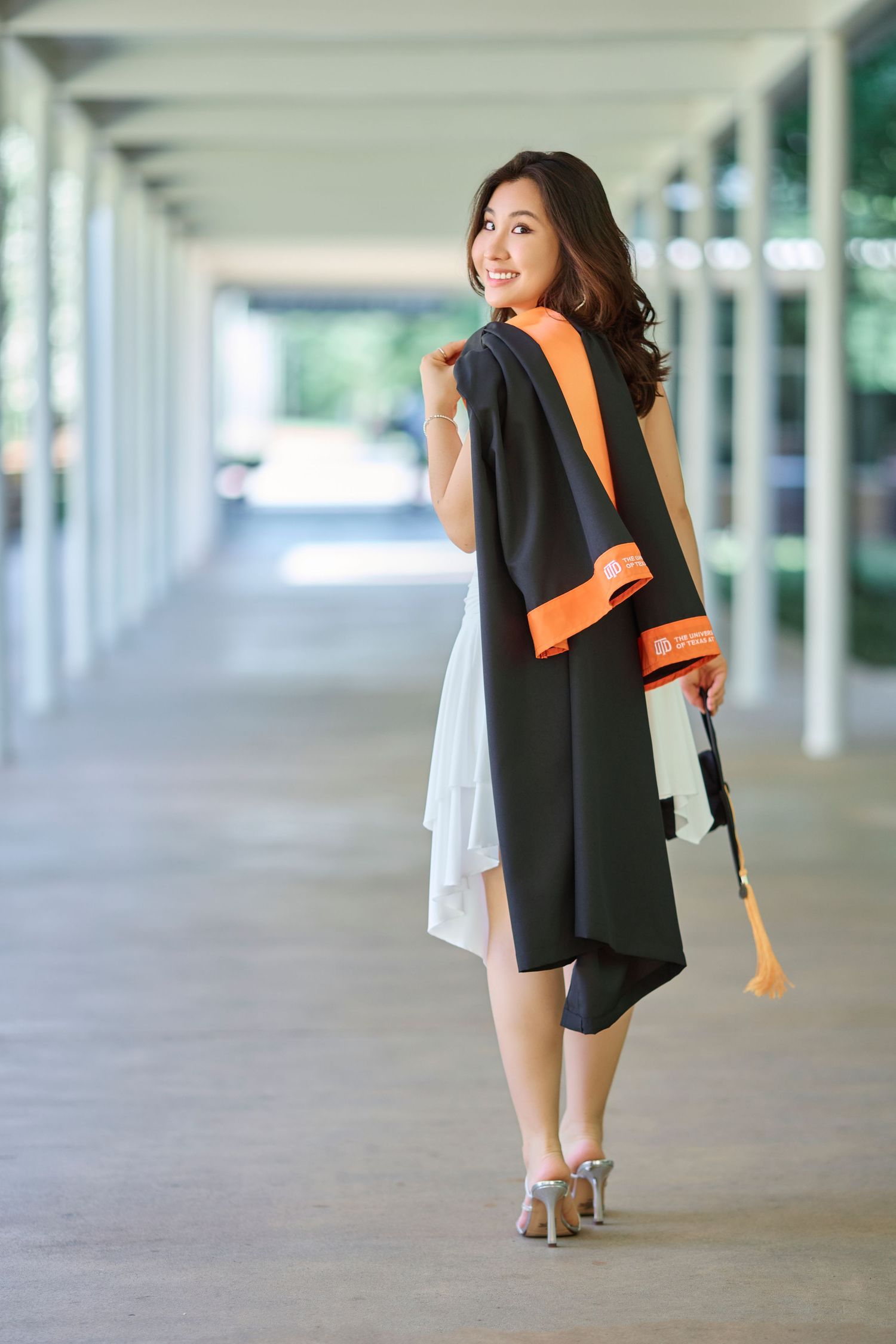 Smiling female graduate in black and orange regalia posing in a sunlit campus corridor.