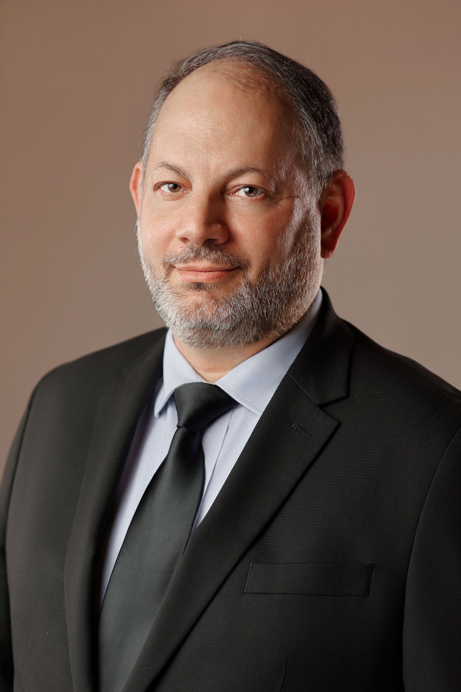 Professional headshot of a middle-aged bearded man in a black suit and tie against a brown background.