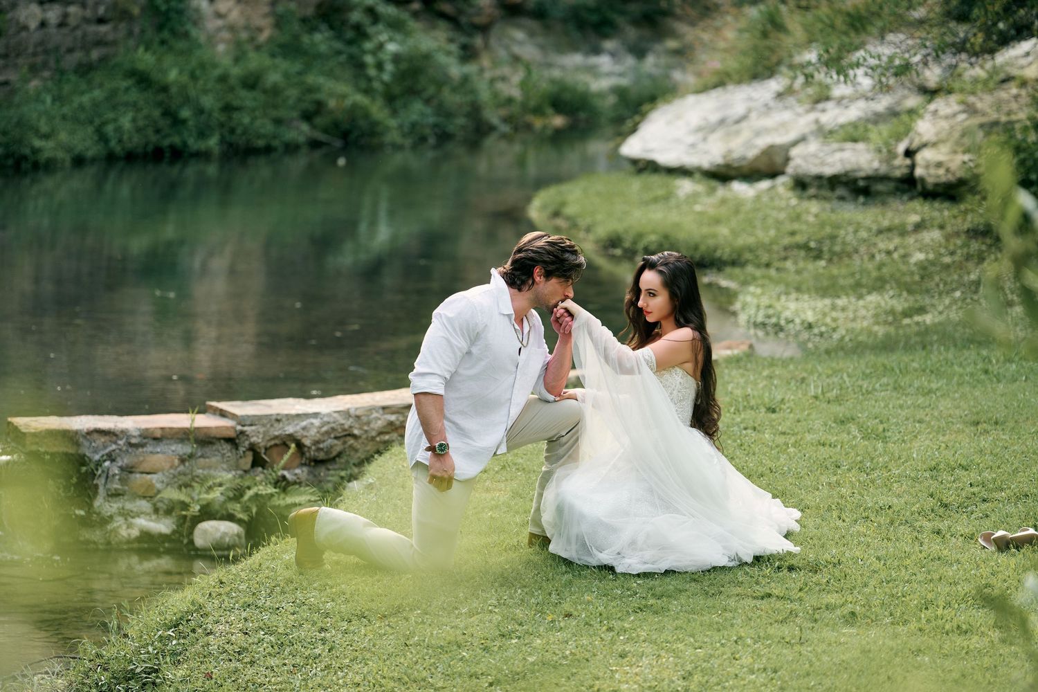 Couple in white attire sharing a romantic moment by a serene river, sitting on lush green grass outdoors.