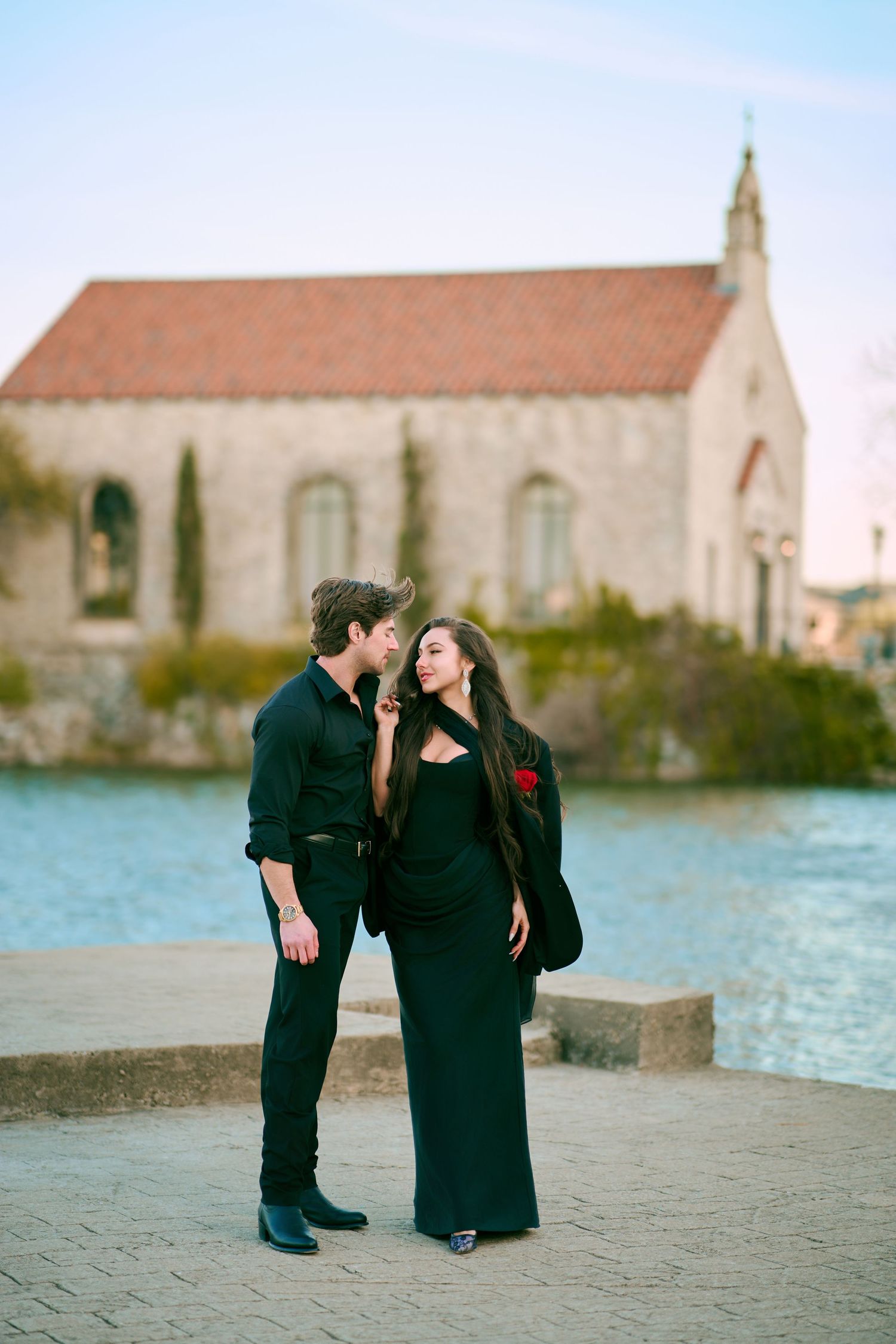 Stylish couple dressed in black standing by waterfront with historic chapel in background.