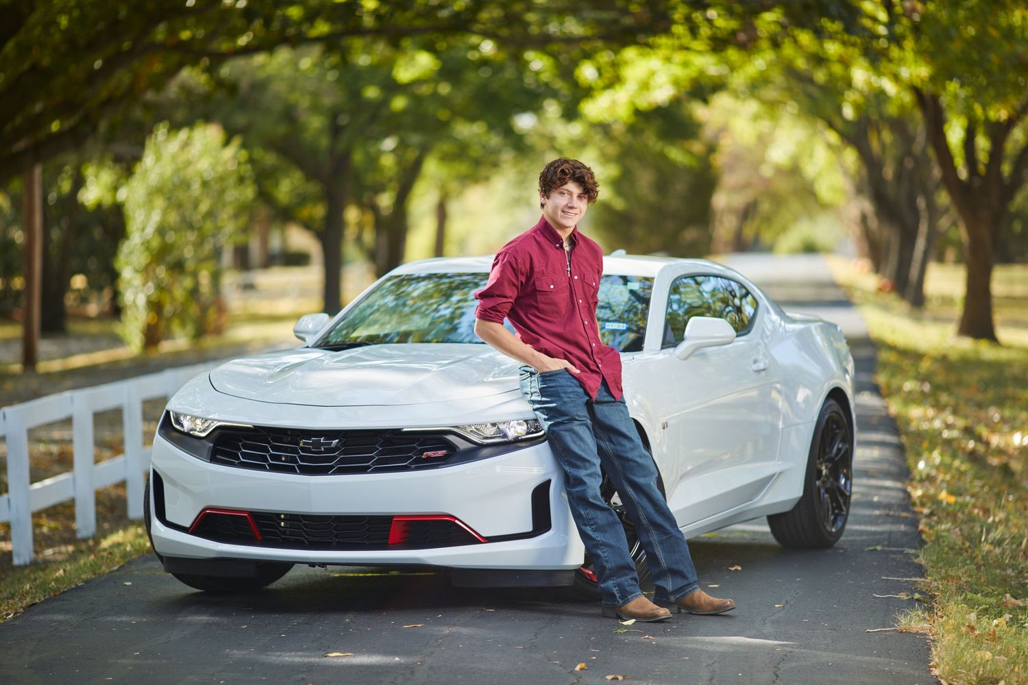 Smiling teenage boy in red shirt leaning against a white Chevrolet Camaro on a tree-lined driveway.