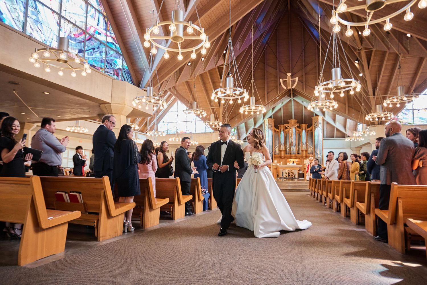 Bride walks down the aisle with escort in elegant church wedding ceremony with wooden pews and chandelier lighting.
