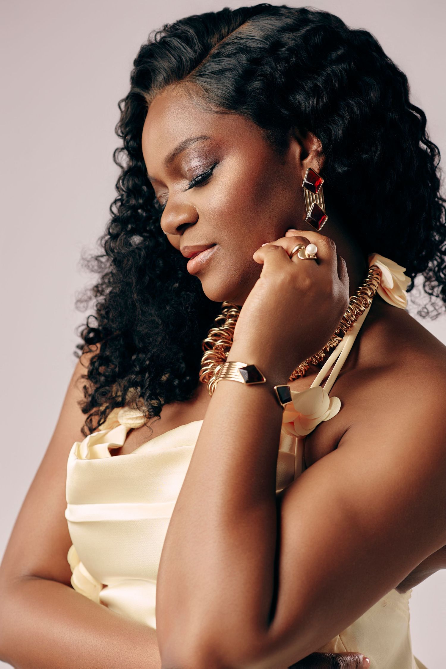 Elegant Black woman in yellow satin dress with gold jewelry, eyes closed, curly hair, studio portrait.