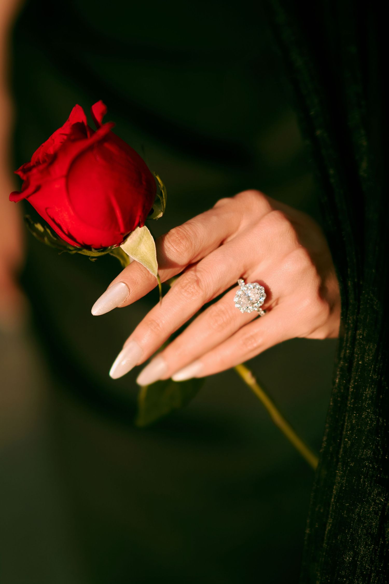 A woman's hand with long nude nails and a diamond ring holds a red rose against a dark green background.