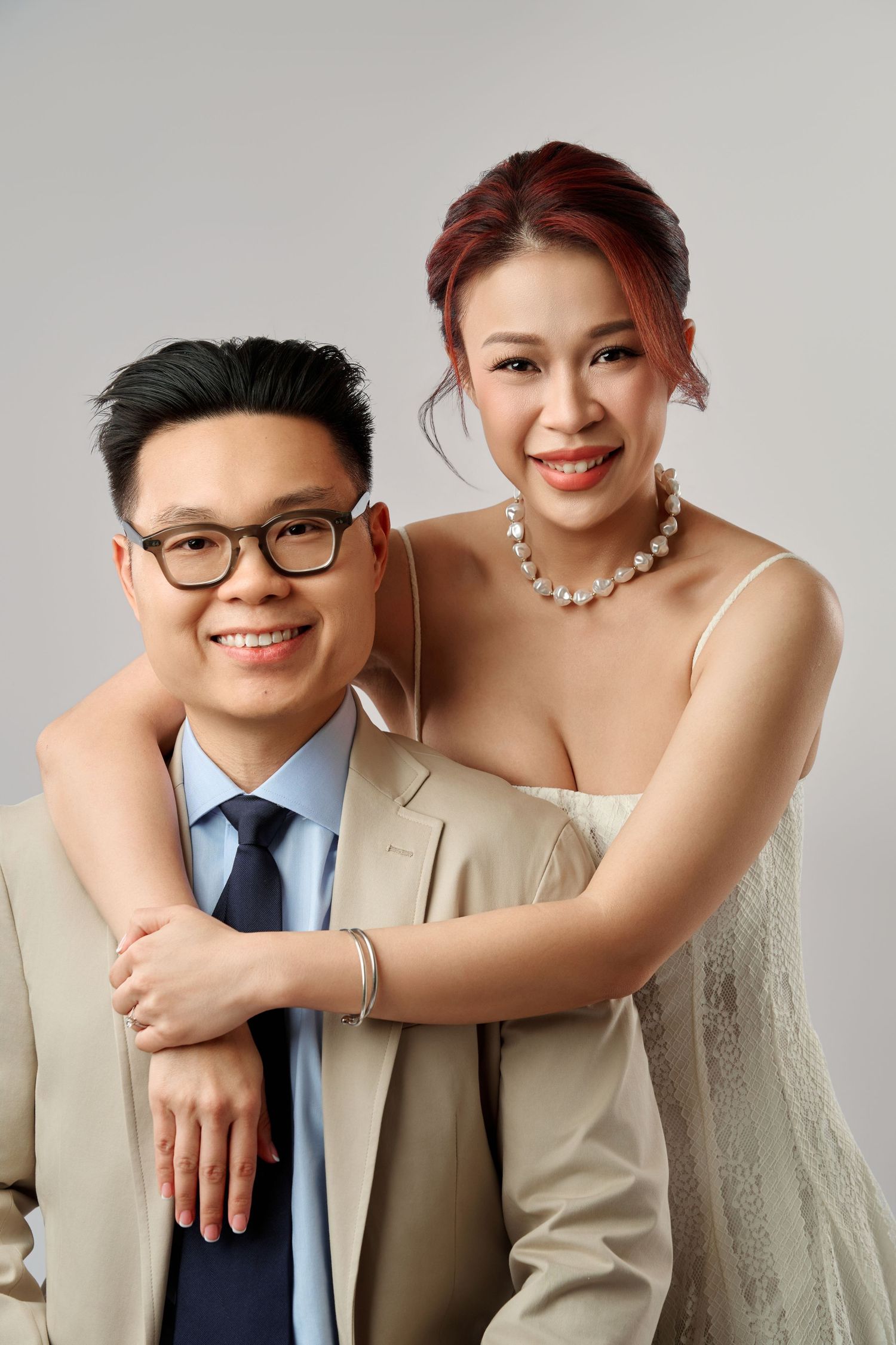 Smiling Asian couple in elegant attire, man in suit with tie and glasses, woman in white dress with pearl necklace.