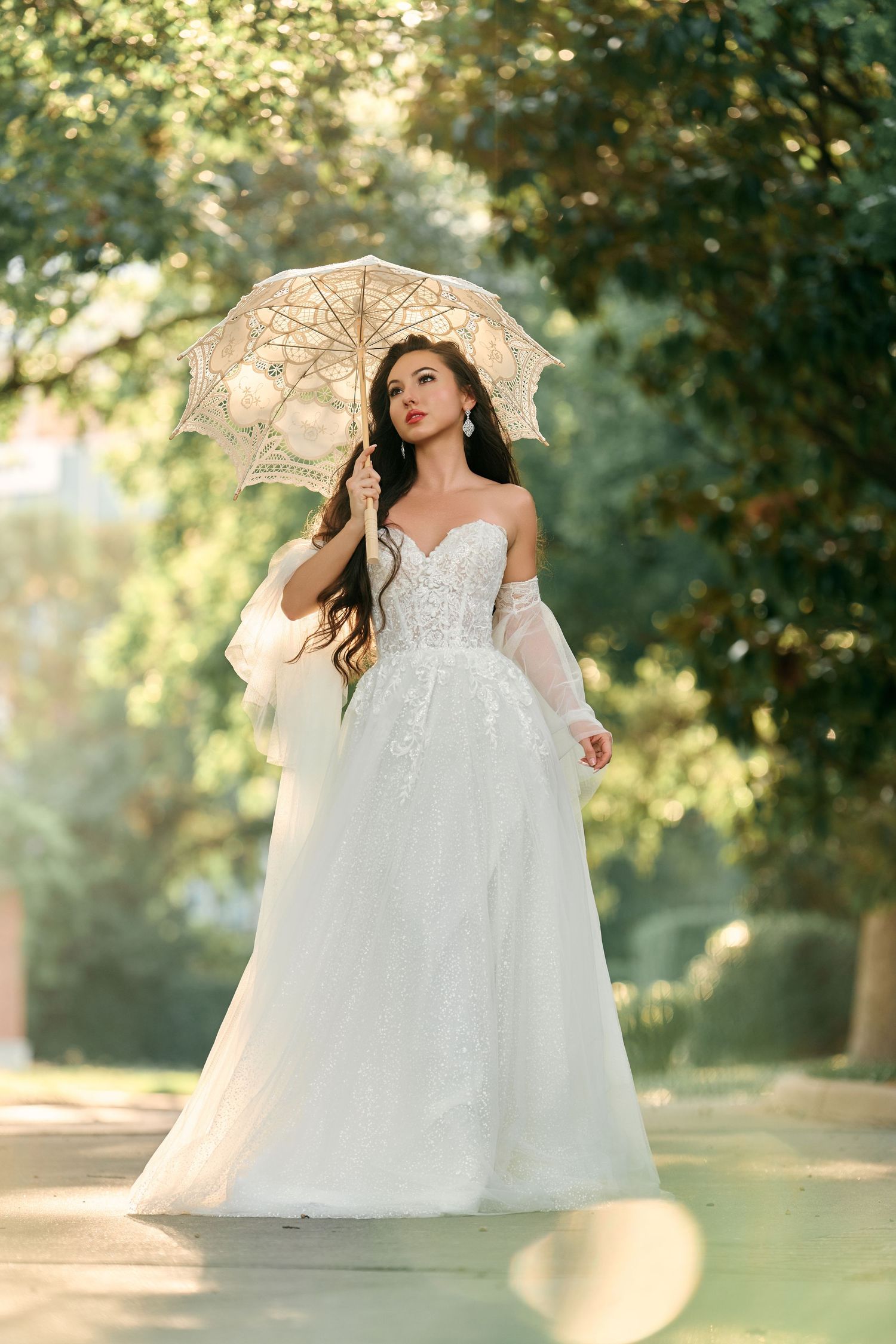 Bride in white lace A-line gown holding vintage parasol umbrella outdoors in sunlit garden setting.