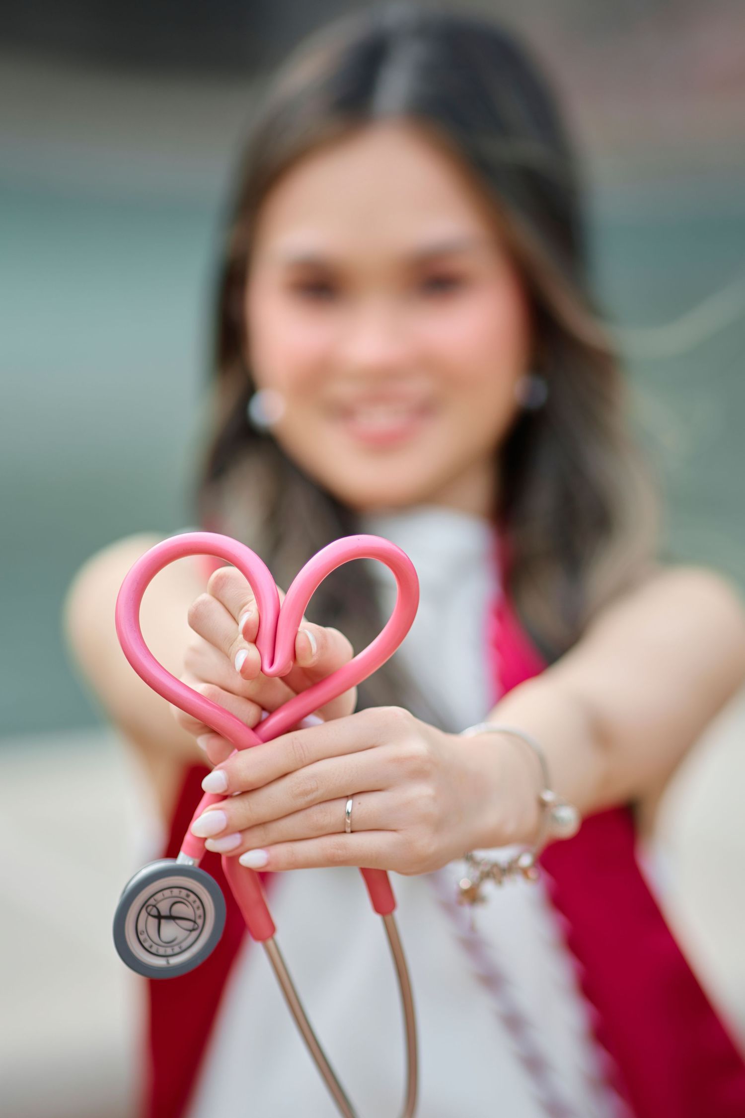Nursing graduate smiles while forming heart shape with pink stethoscope toward camera.