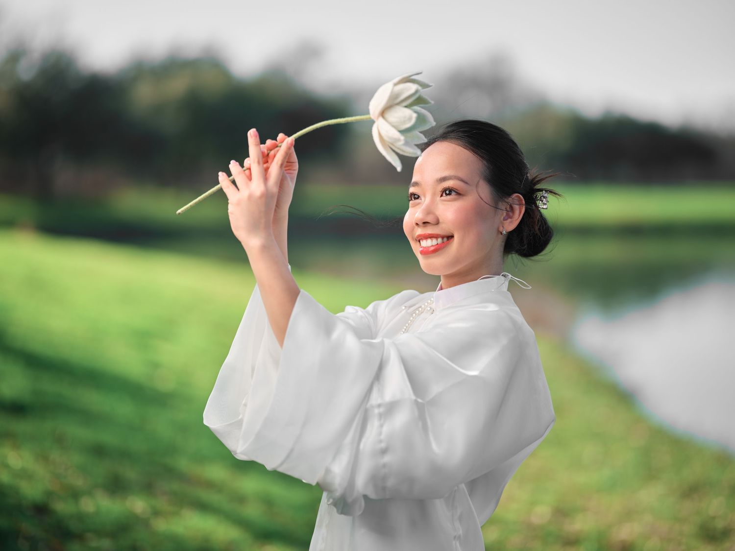 Smiling Asian woman in white flowing outfit tossing a lotus flower outdoors near a peaceful pond.