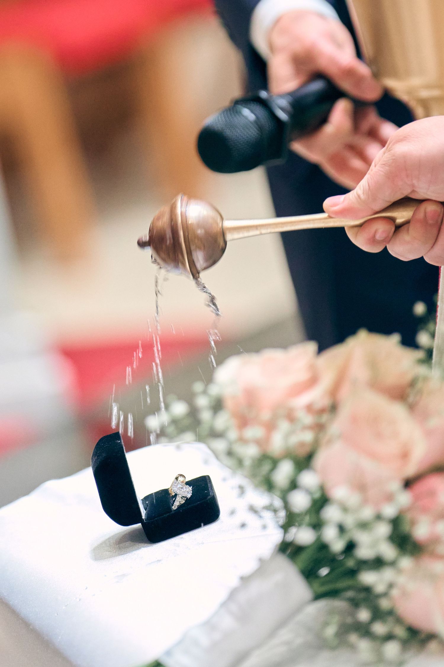 A priest sprinkles holy water over wedding rings in a velvet box during a religious ceremony with roses nearby.