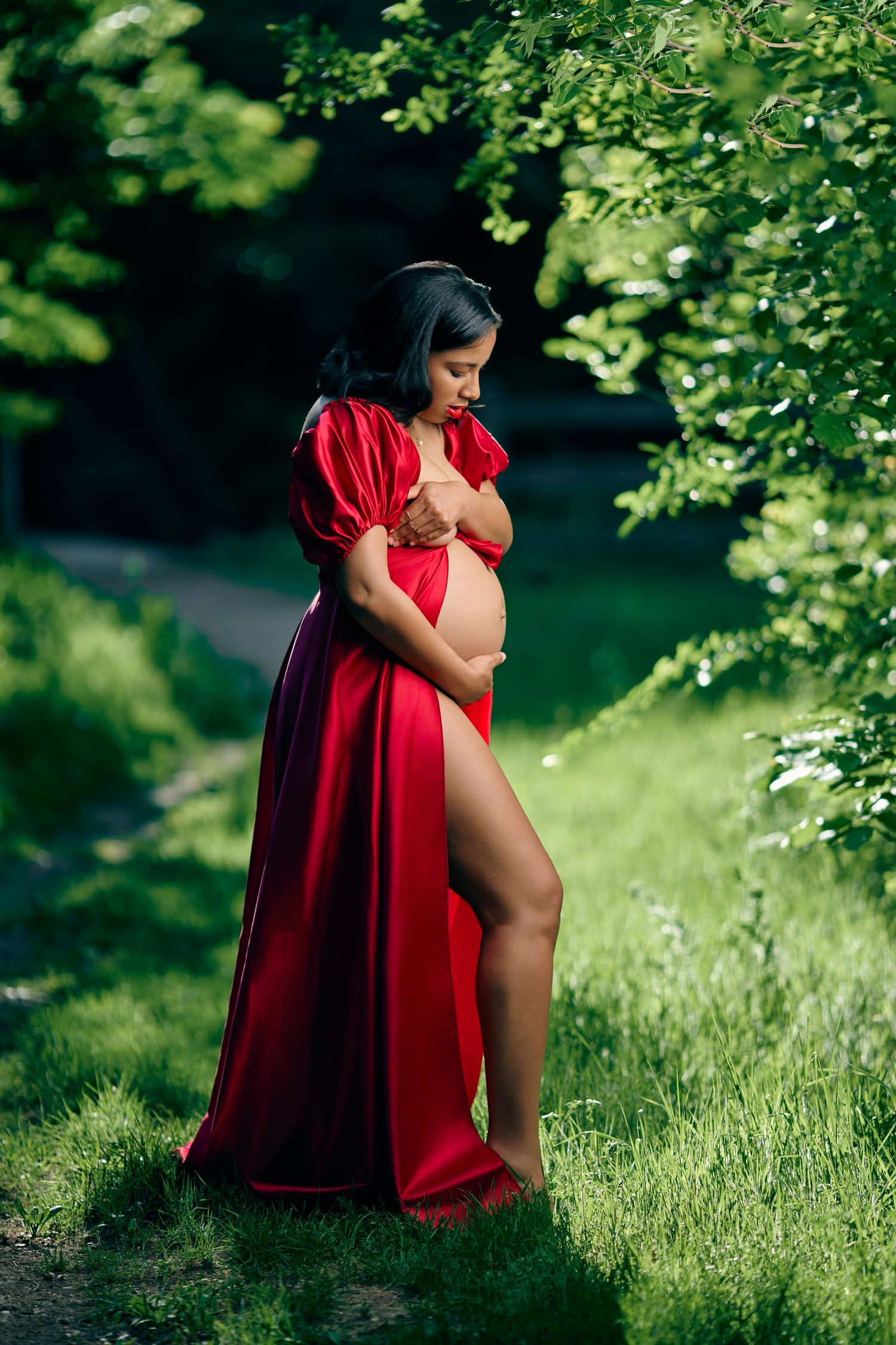 Pregnant woman in a flowing red satin gown with a thigh-high slit standing in a lush green forest.